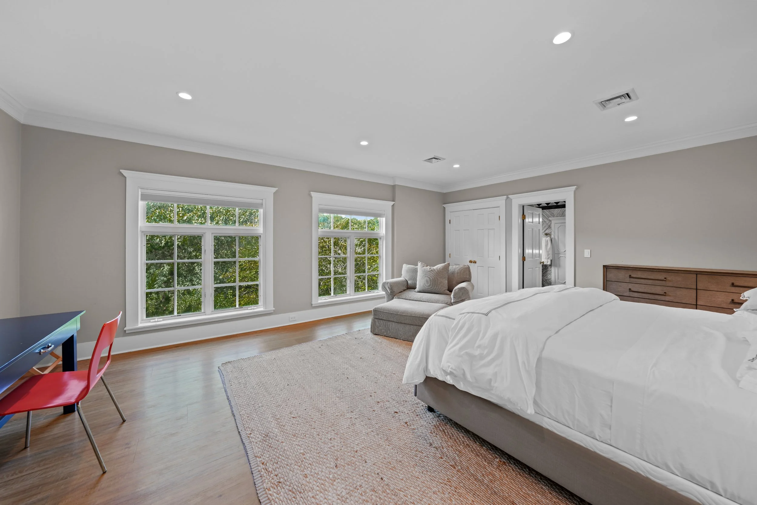 A spacious bedroom with light grey walls, large windows with white trim, a bed with white bedding, a wooden dresser, a grey armchair with pillows, a small black desk with a red chair, and a beige area rug on a hardwood floor.