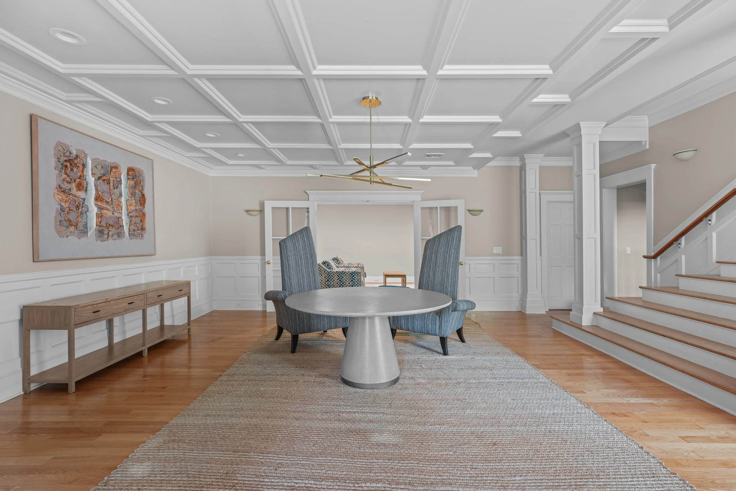 A spacious dining room with a round table, attached with four high-back gray chairs, on a beige and brown striped rug. Behind the table is a large window with white frames, flanked by two green wall sconces. To the left is a wooden console table agai