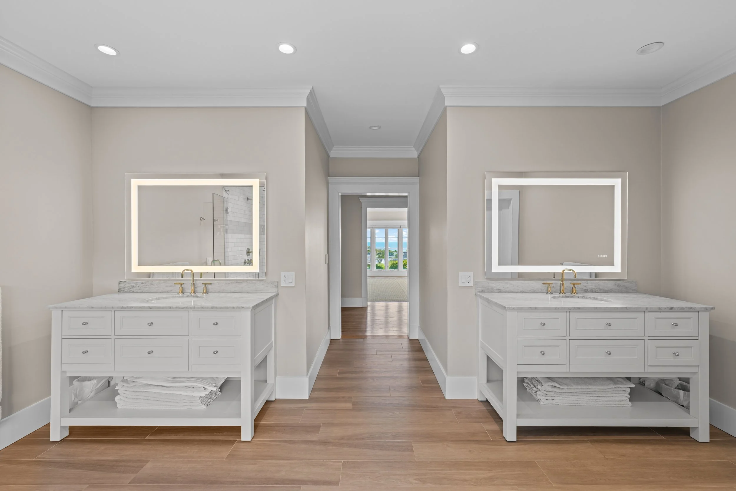 Two identical white bathroom vanities with marble countertops, gold faucets, large rectangular mirrors with built-in lighting, and neatly folded towels underneath. The vanities are separated by a hallway leading to a bright room with windows.