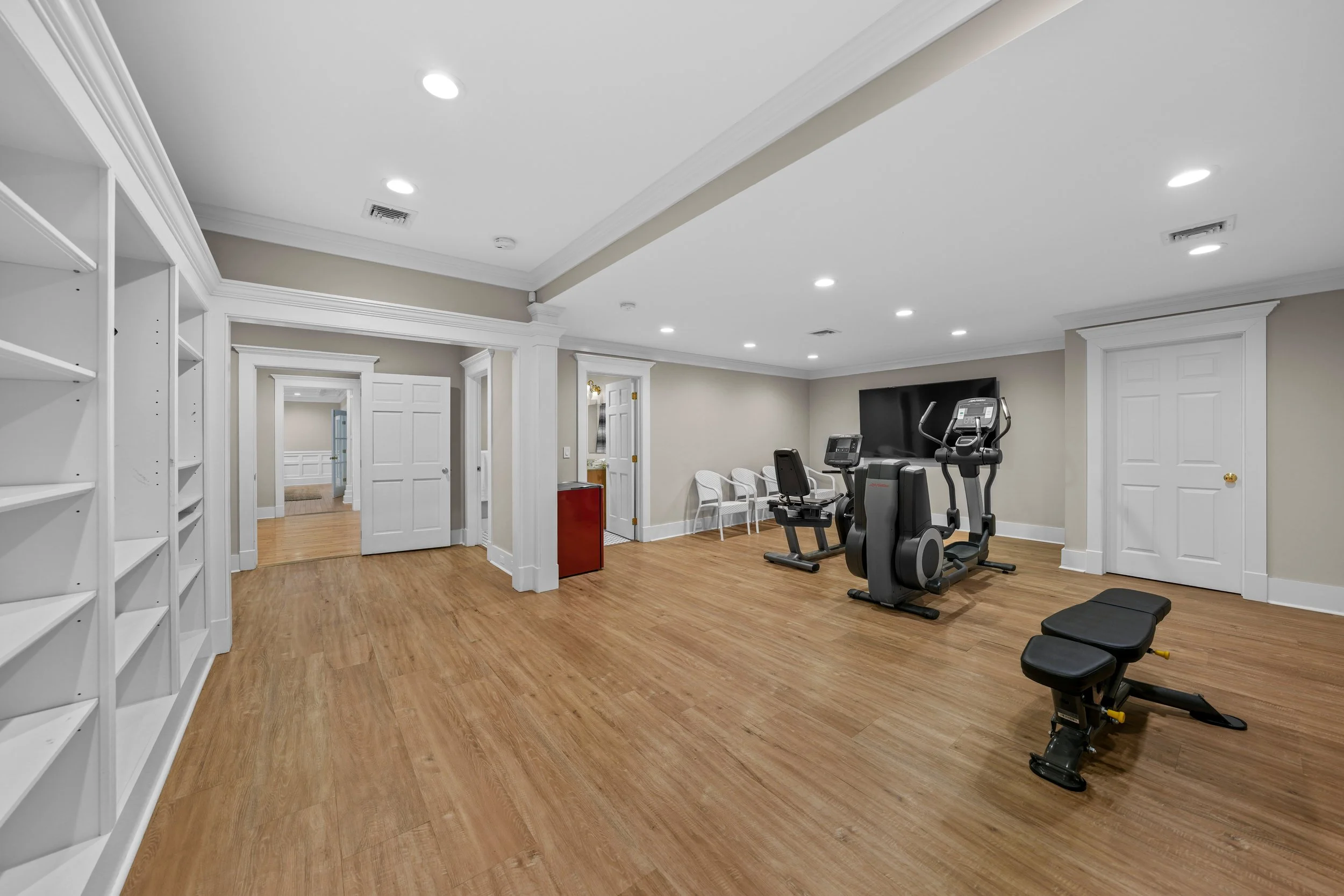 Home gym with cardio equipment, benches, and white chairs, hardwood floors, and recessed ceiling lights.