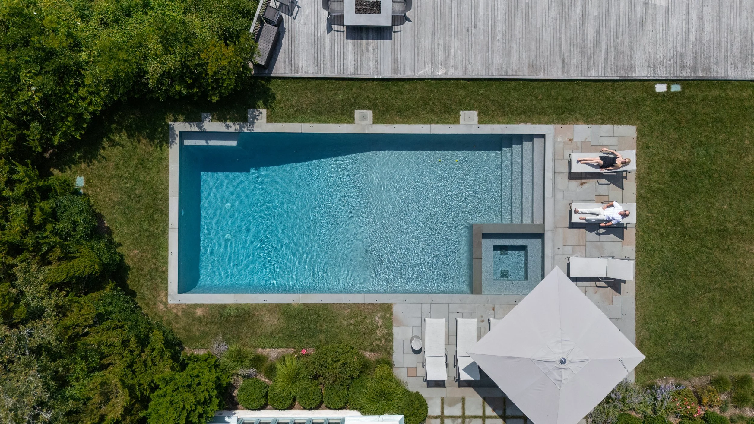 Aerial view of a backyard swimming pool with surrounding grass, patio furniture, and two people lounging on chairs under a white umbrella.