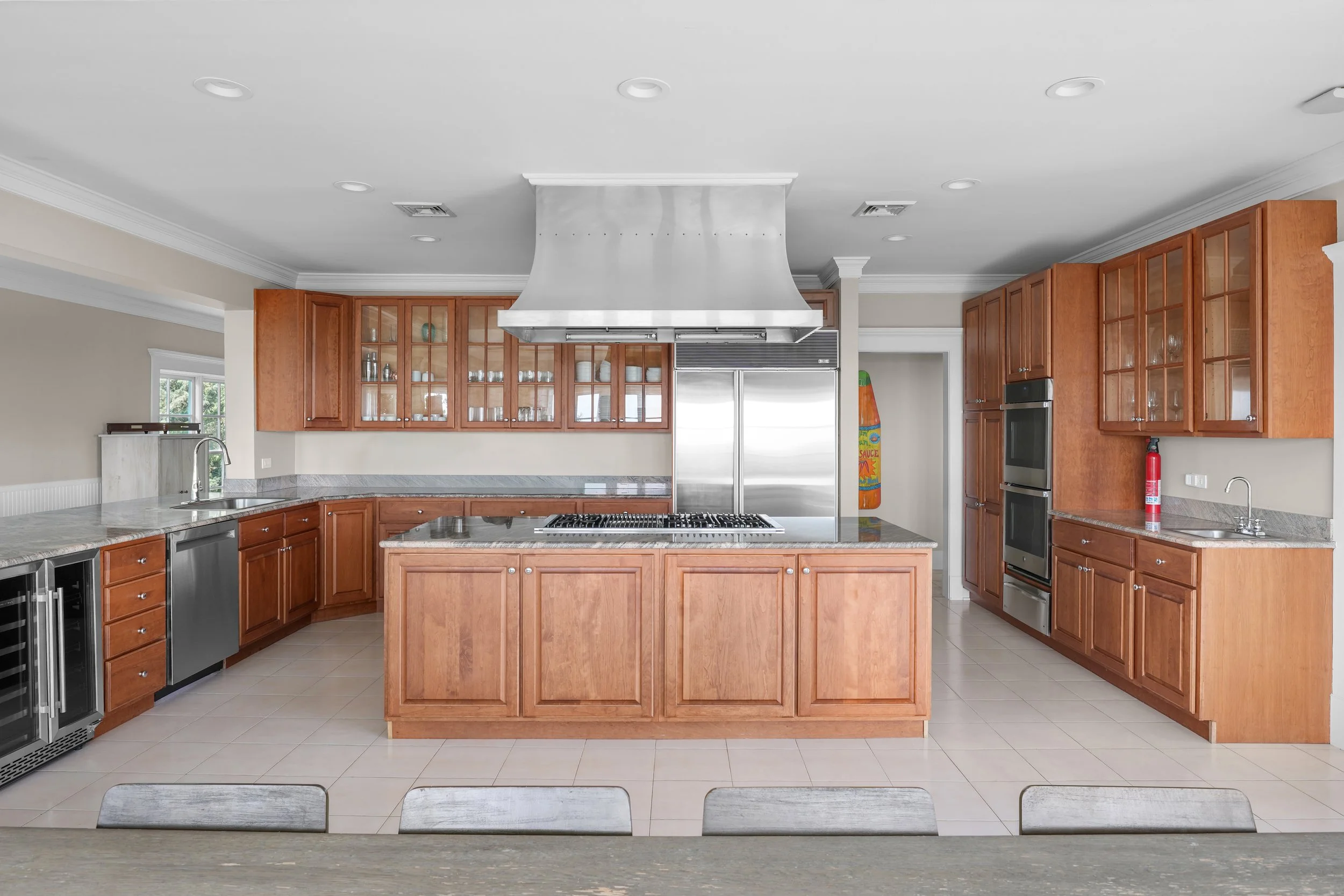 Modern kitchen with wooden cabinetry, stainless steel appliances, and a central island with a cooktop, under a large stainless steel exhaust hood.