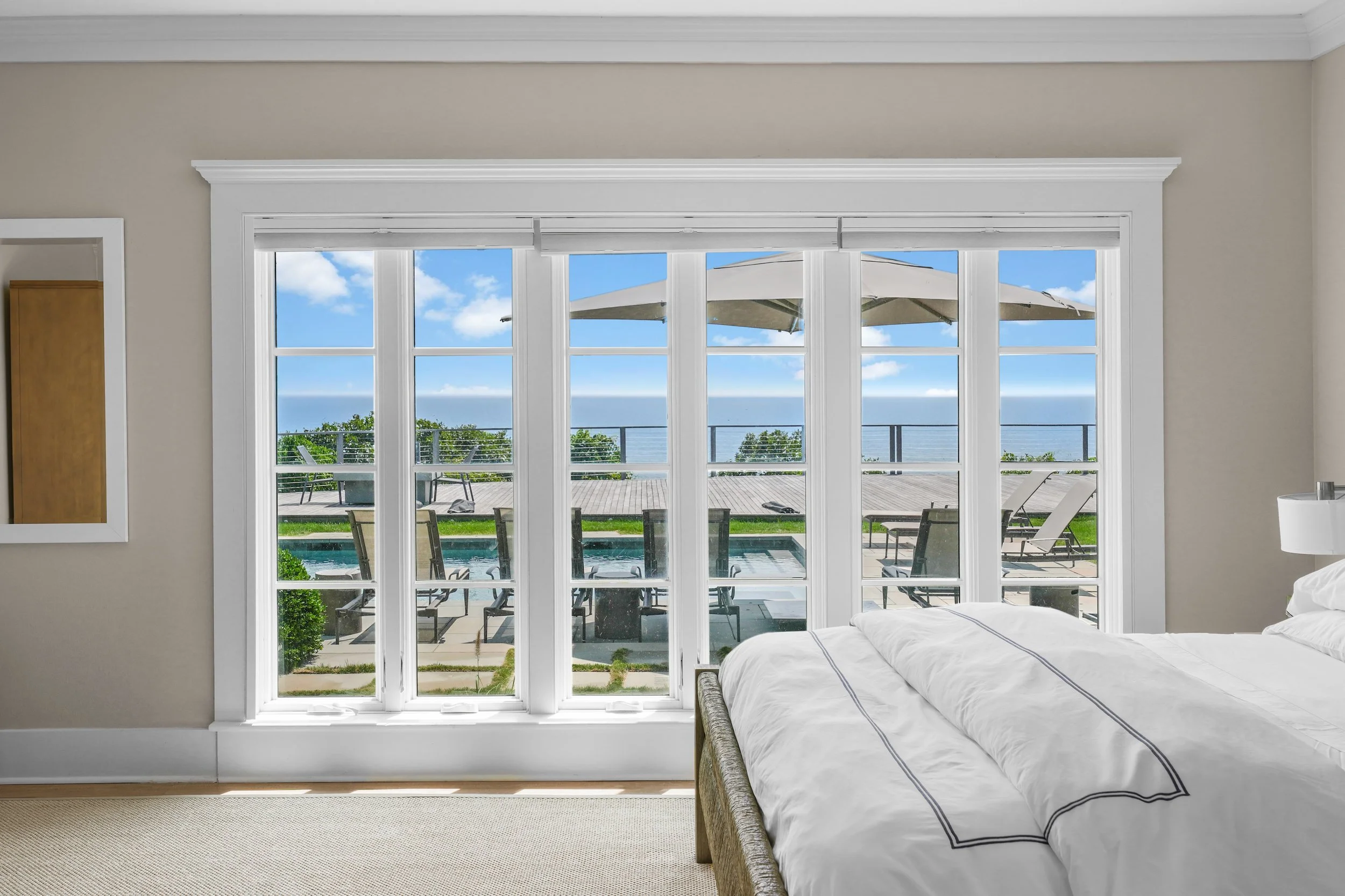View of a bedroom with a large window overlooking a patio, swimming pool, and ocean in the distance, with chairs and an umbrella outside.