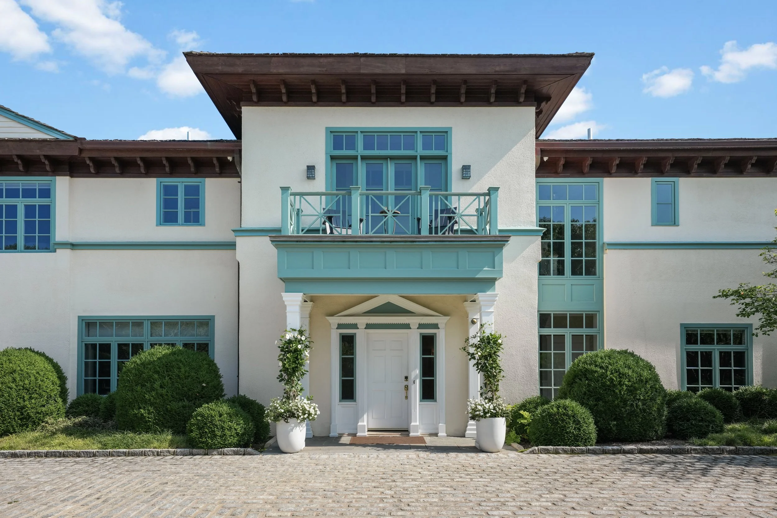 Front view of a modern, white two-story house with teal window frames, a teal balcony, and decorative teal accents. The house features white columns at the entrance, surrounded by potted plants, and lush bushes in the front yard. The sky is blue with