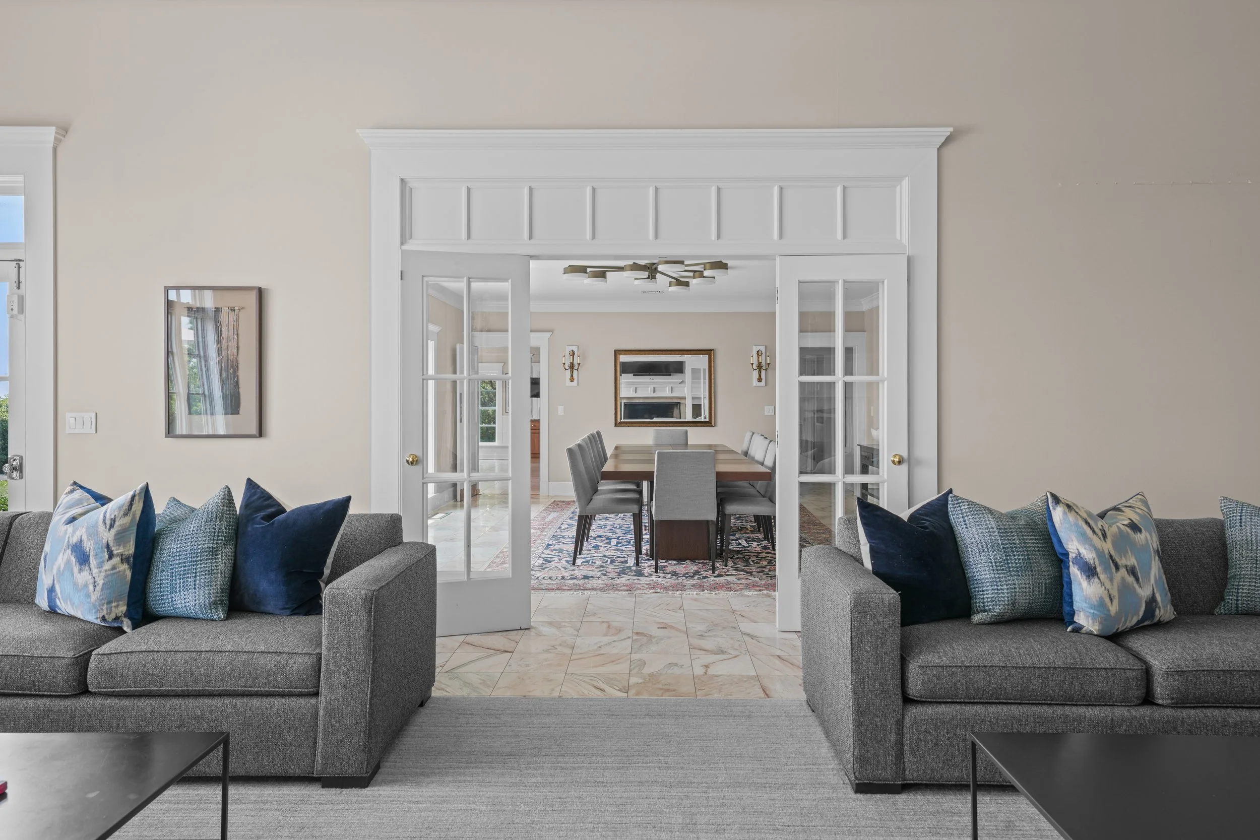 Living room with gray sofas and pillows facing a dining room with a wooden table, eight gray chairs, and a mirror on the wall, viewed through white double French doors.