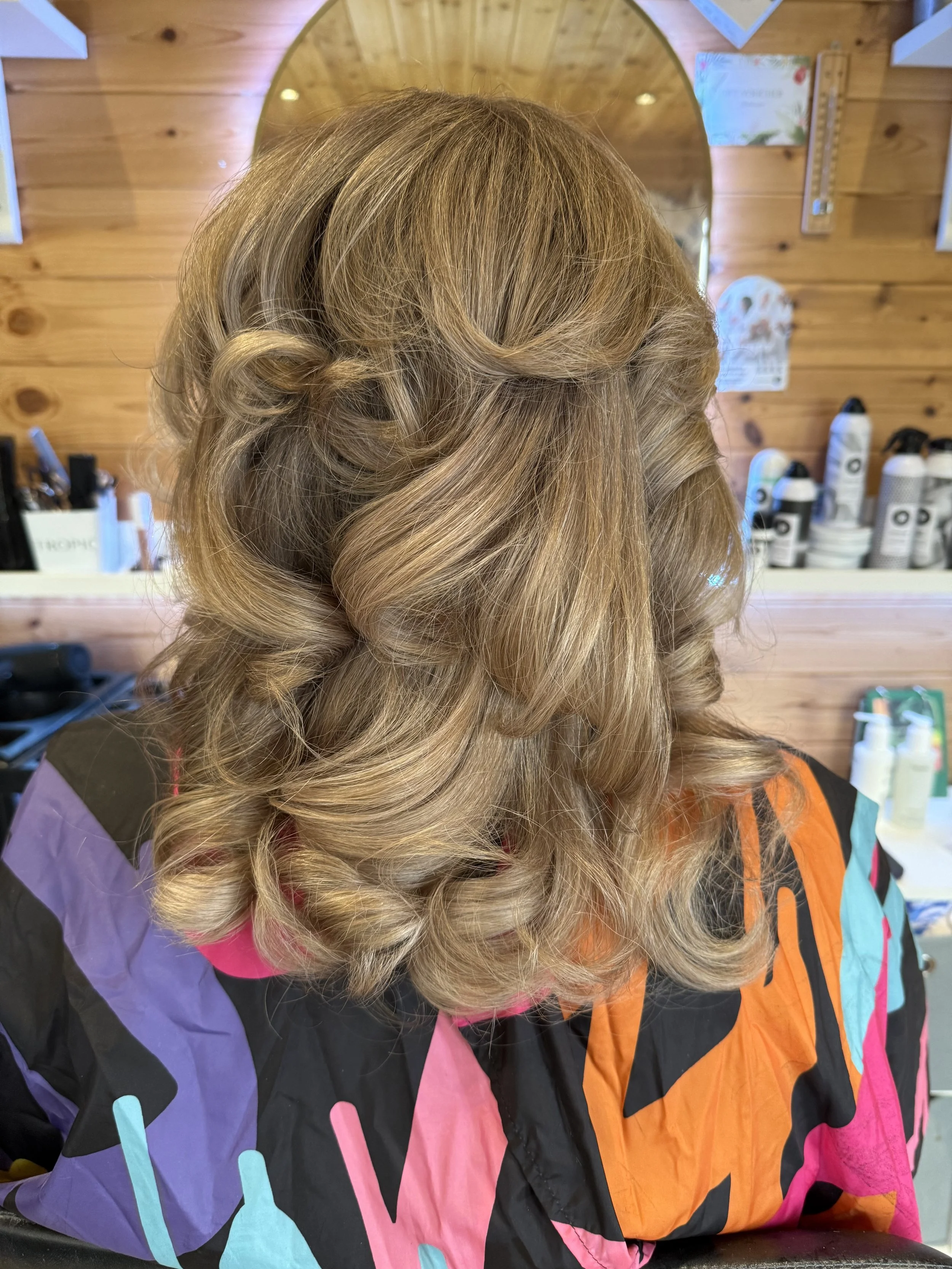 A woman's head with styled, shoulder-length, wavy blonde hair. She is seated in a salon with wooden walls and bottles of hair products visible in the background.