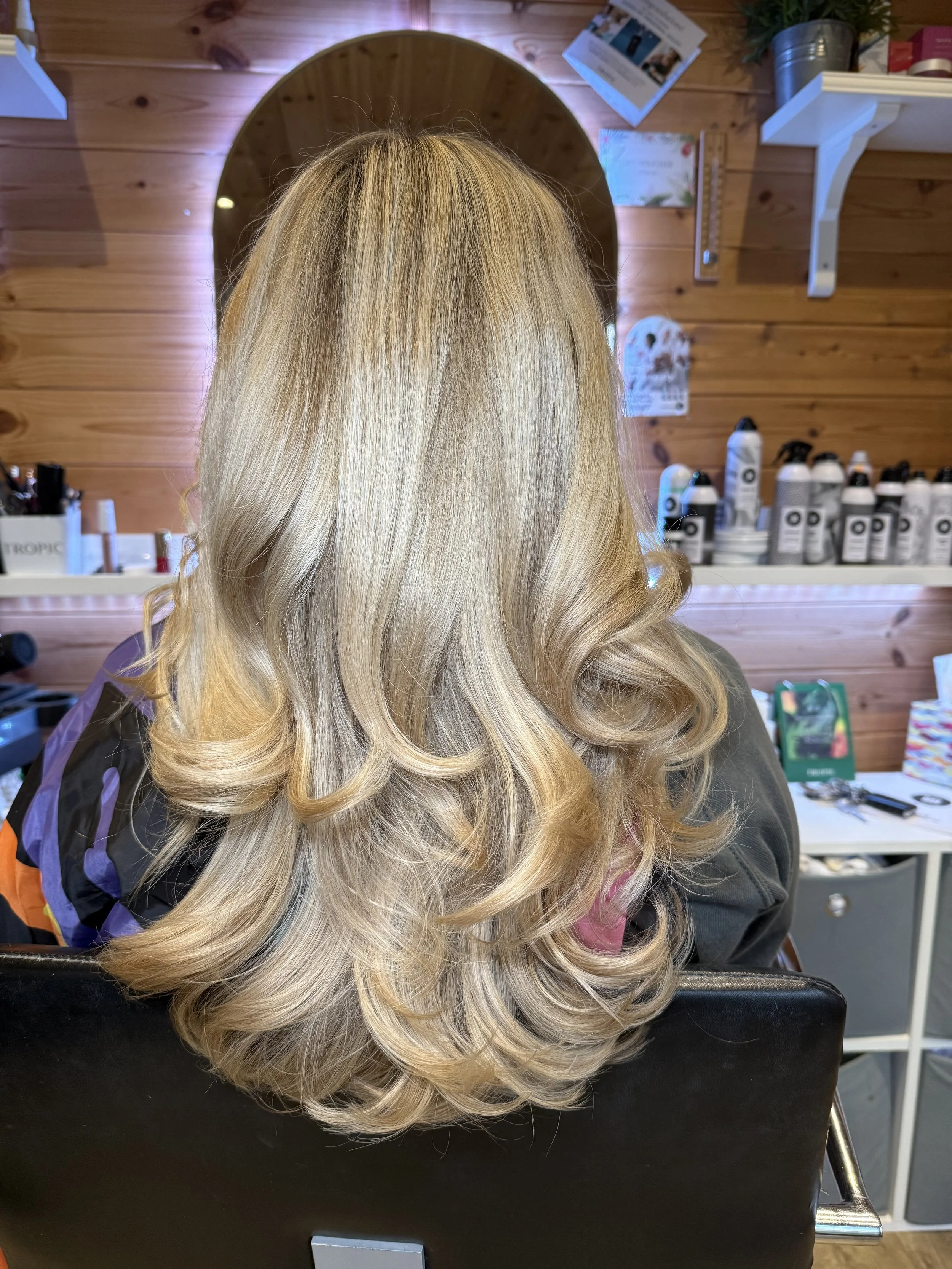 Back view of a woman with long, blonde, styled hair with loose curls, sitting in a salon with a wooden wall and shelves with hair products in the background.