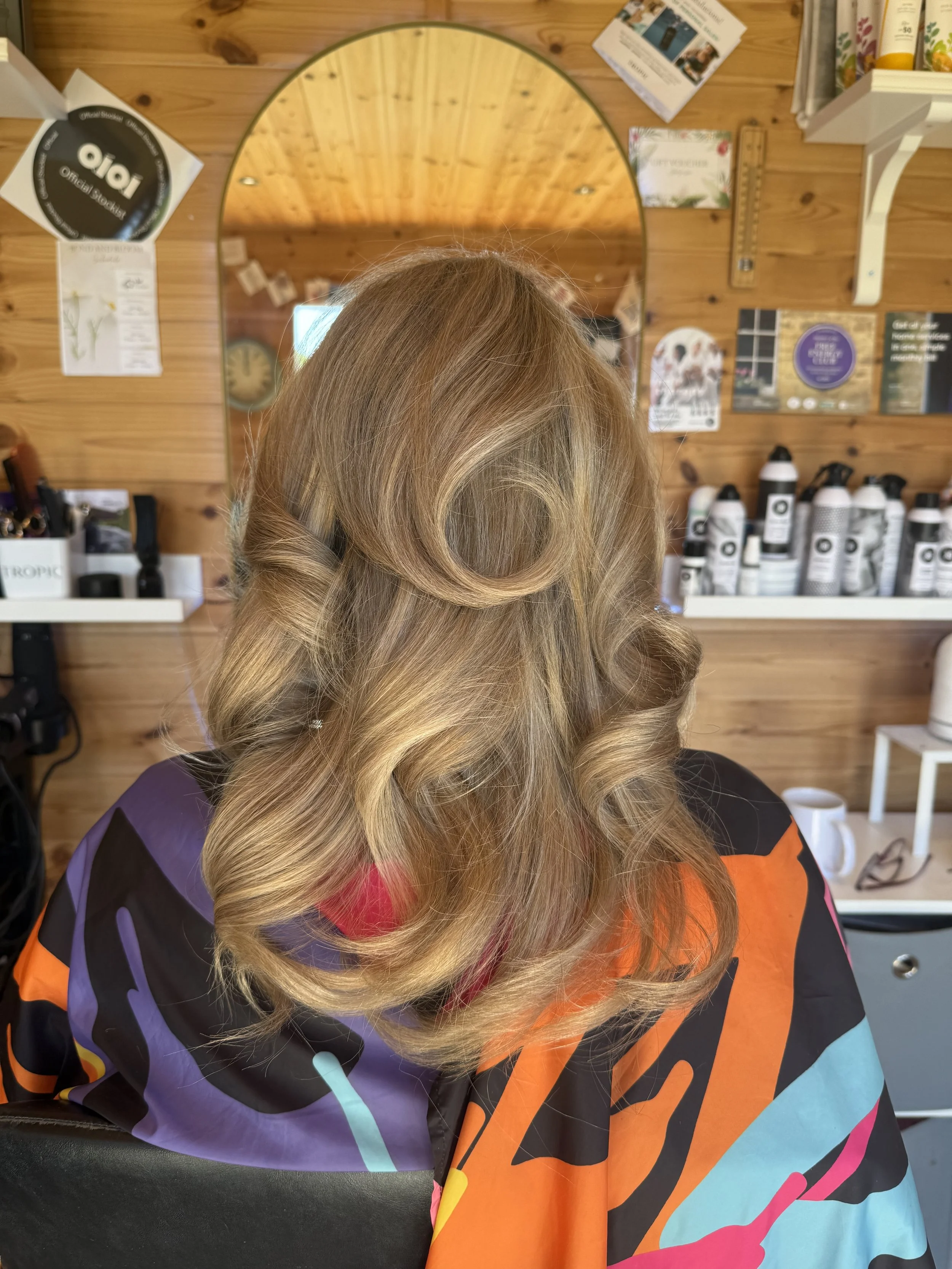 A woman with styled, blonde hair sitting in a salon with a wooden wall, mirror, and hair products in the background.