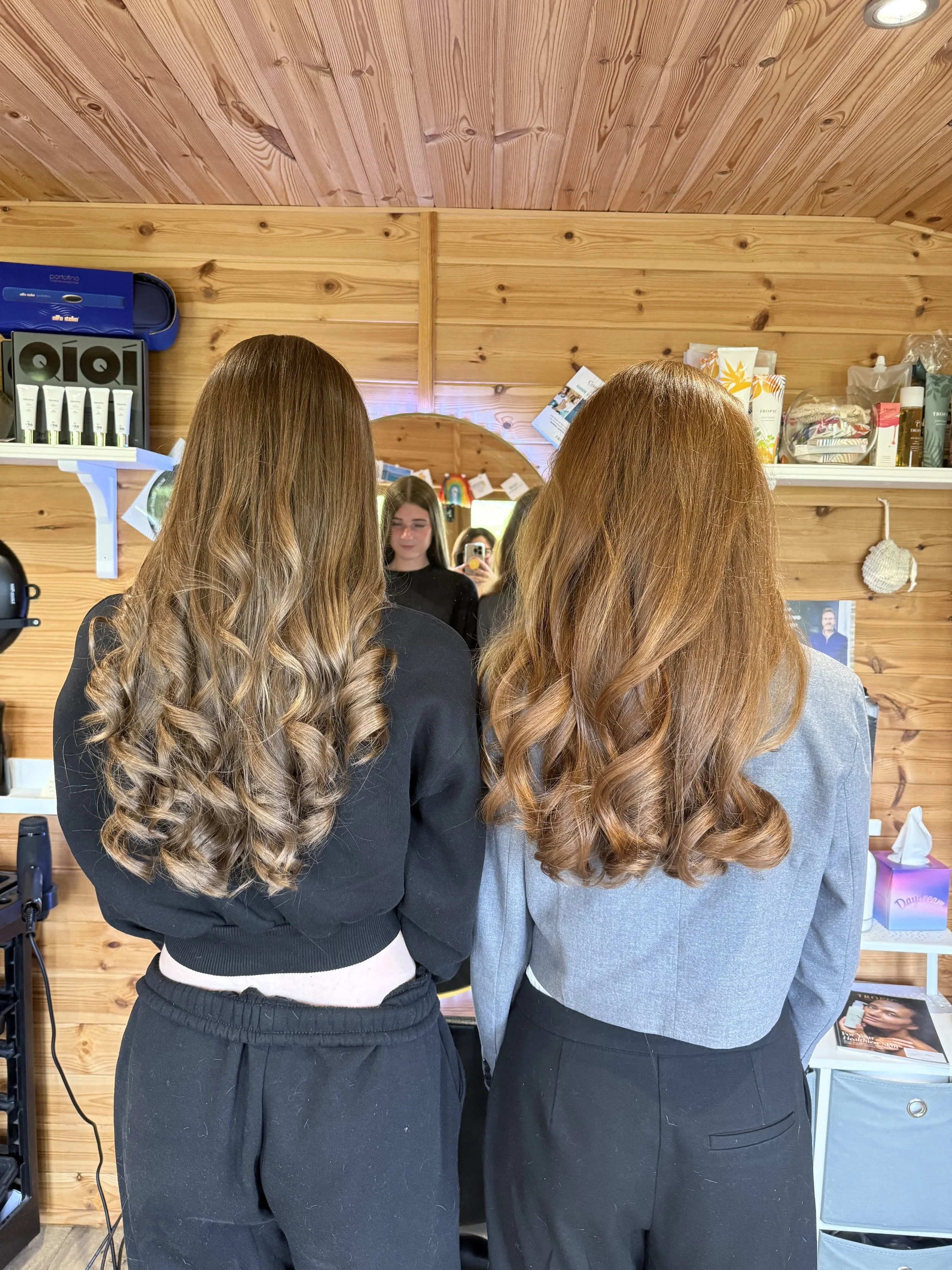 Two women with long, wavy brown hair standing side by side in front of a mirror in a wooden-paneled room, with a third woman taking a photo of their reflection.