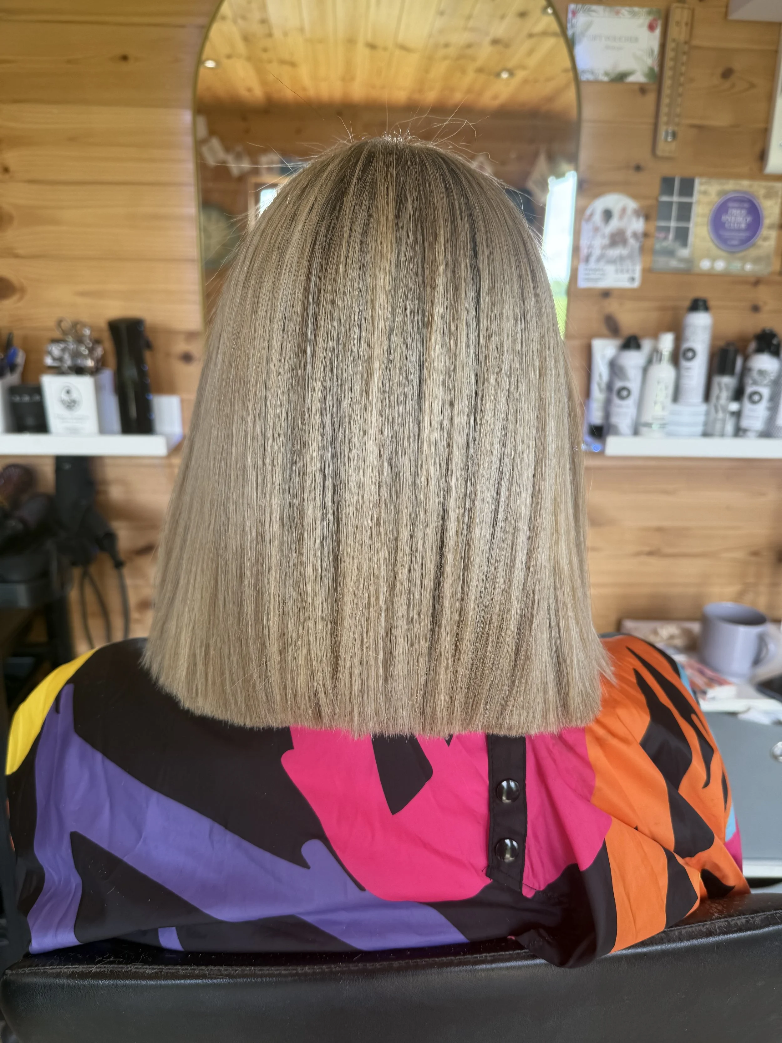 The image shows the back of a person's head with a straight, blonde bob haircut. They are seated in a barbershop or salon with wooden walls and various hair products visible on shelves in the background.