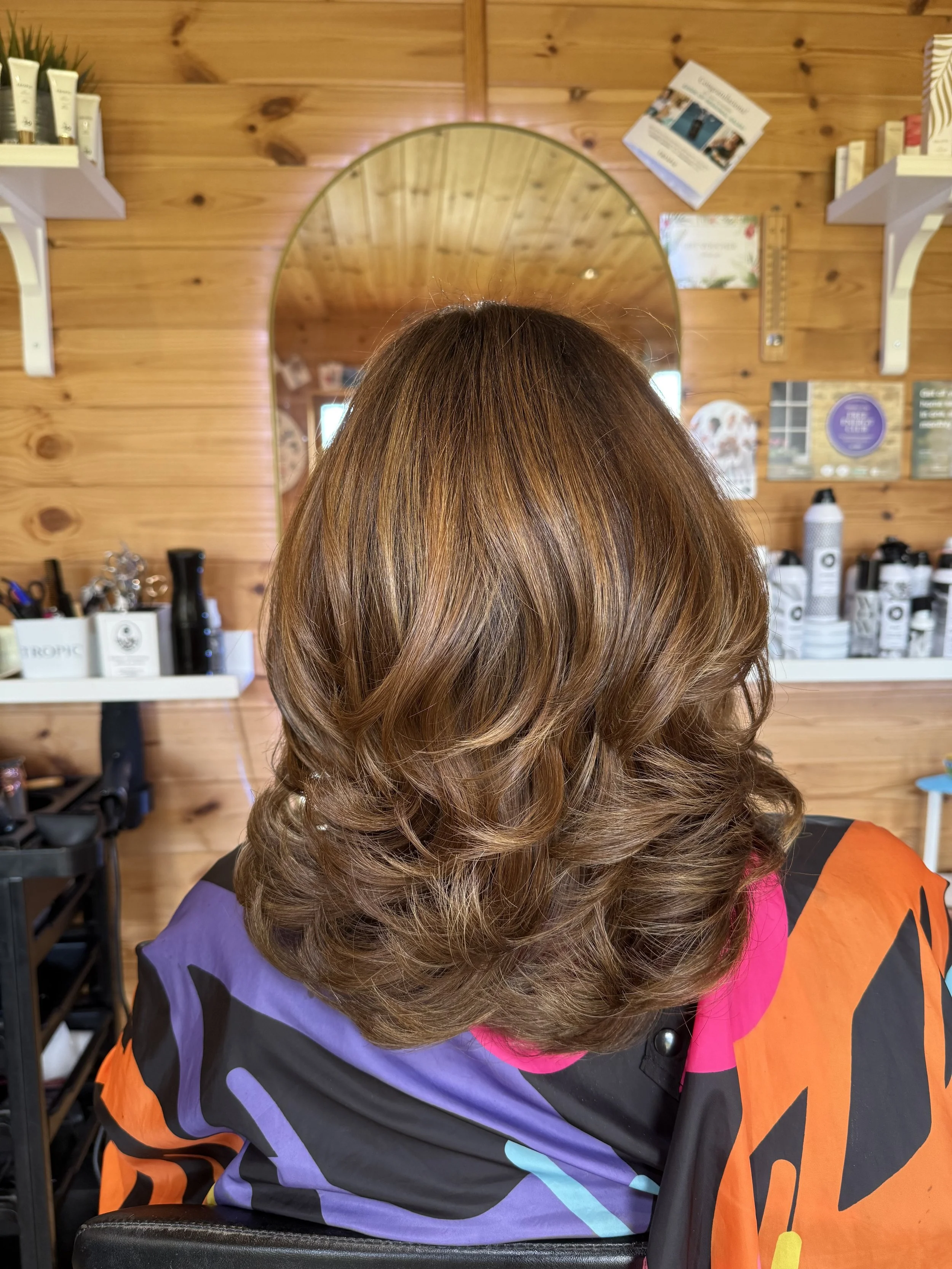 The back of a woman's head with styled, wavy brown hair, sitting in a hair salon with wooden walls, mirror, and hair products in the background.