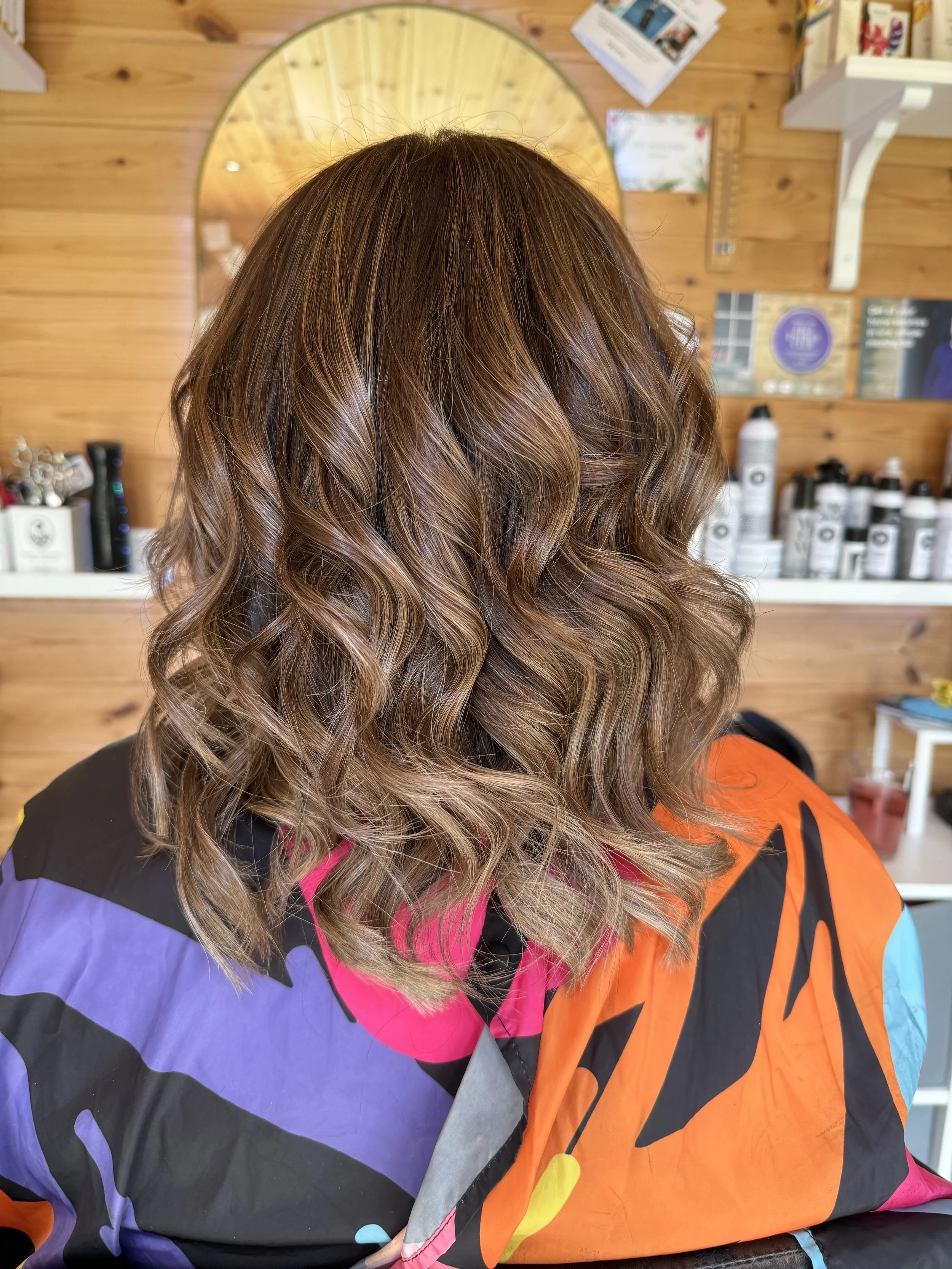 Back view of a woman with shoulder-length, wavy, light brown hair sitting in a hair salon with wooden walls and shelves with hair products in the background.