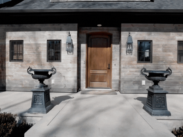 Front view of a house entrance with a wooden door, two black urn planters, and two wall-mounted lanterns on a brick and stone exterior wall.