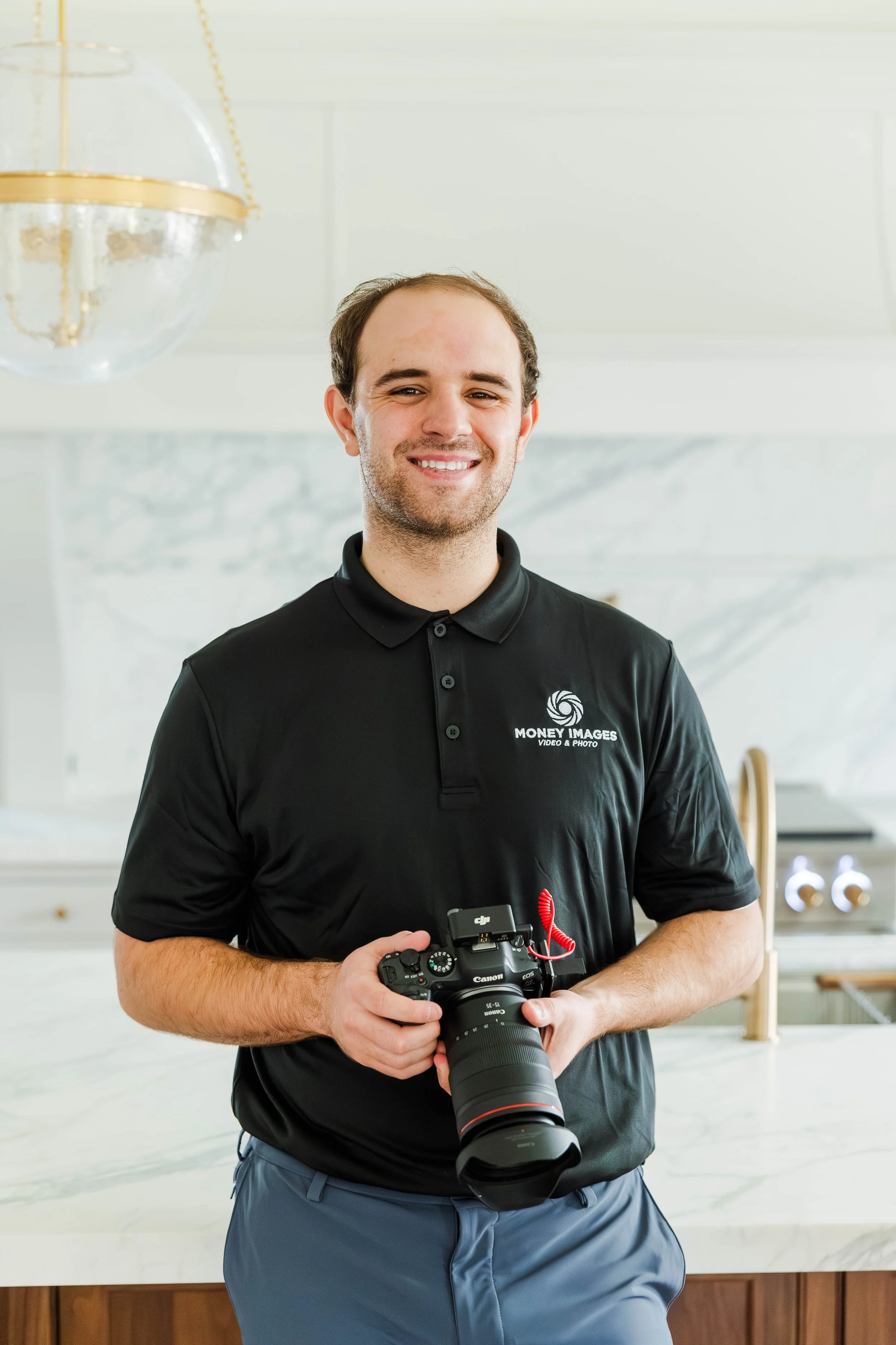 A man smiling and holding a Canon camera while standing in a bright room with white walls and a marble countertop.