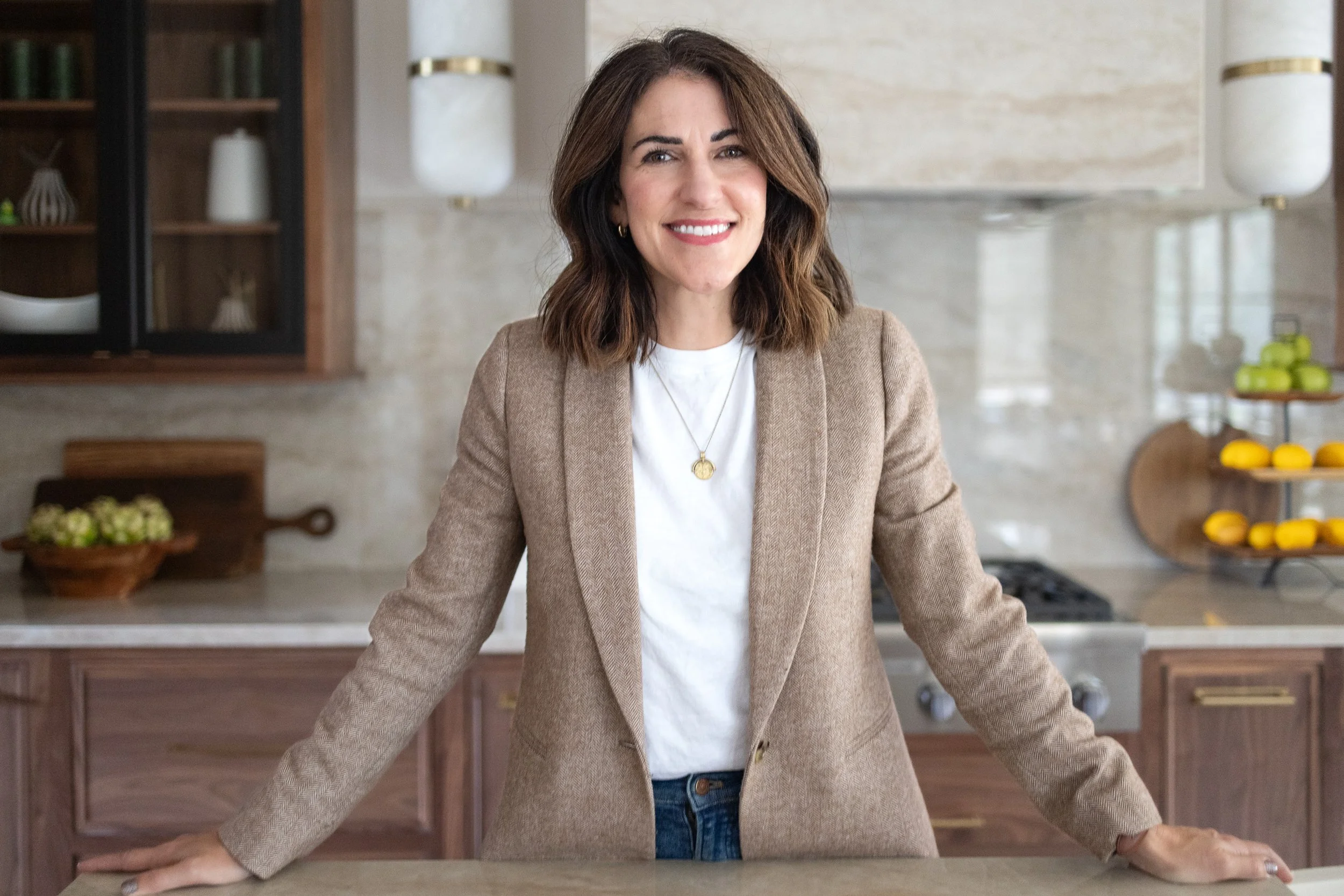 A woman with shoulder-length brown hair and a big smile standing in a modern kitchen, leaning on the counter with both arms, wearing a beige blazer over a white shirt, with a necklace. The kitchen has wooden cabinets, shelves with decorative items, and a light-colored marble backsplash.