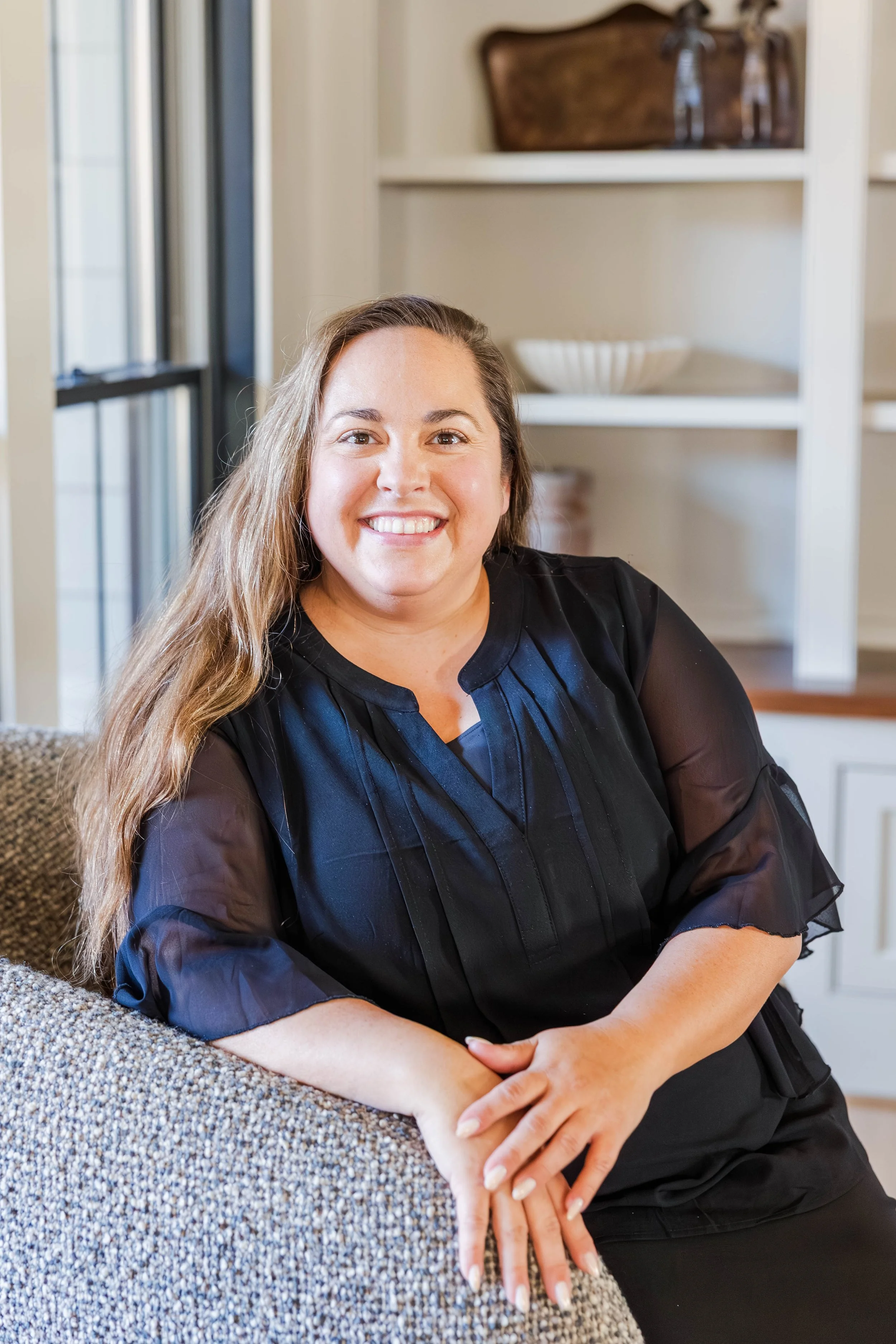 A smiling woman with long, wavy brown hair and light skin, wearing a black dress with sheer sleeves, sitting on a sofa in a cozy home setting with open shelves in the background.