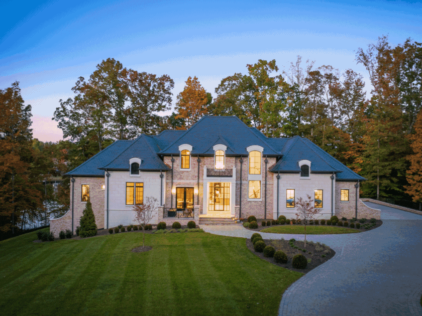 Large luxury house with a blue roof, brick and white exterior, surrounded by green lawn and trees, illuminated from within, during dusk.