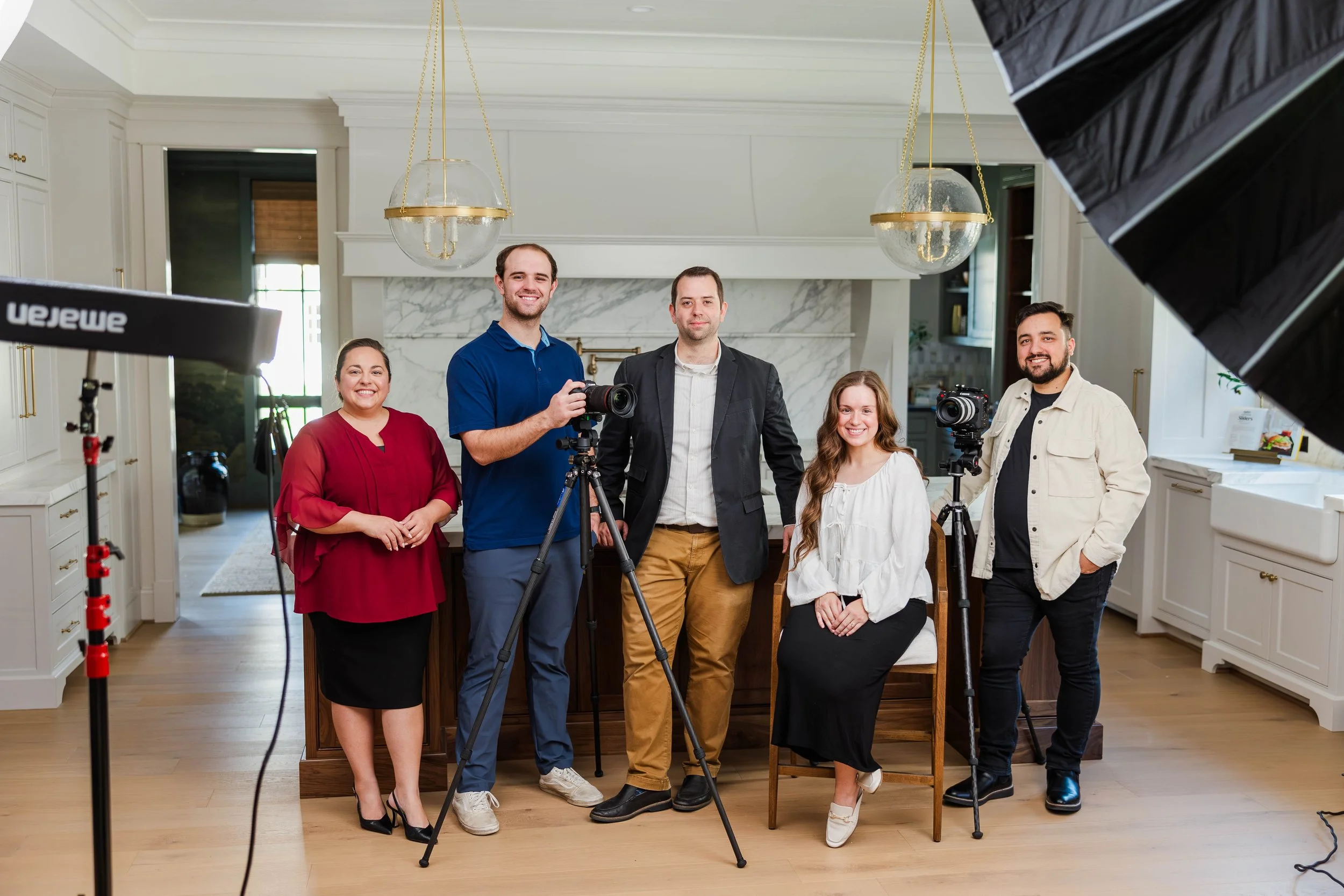 Group of five people posing for a photo in a well-lit modern kitchen, with professional photography equipment including a camera, tripod, and studio lights visible.