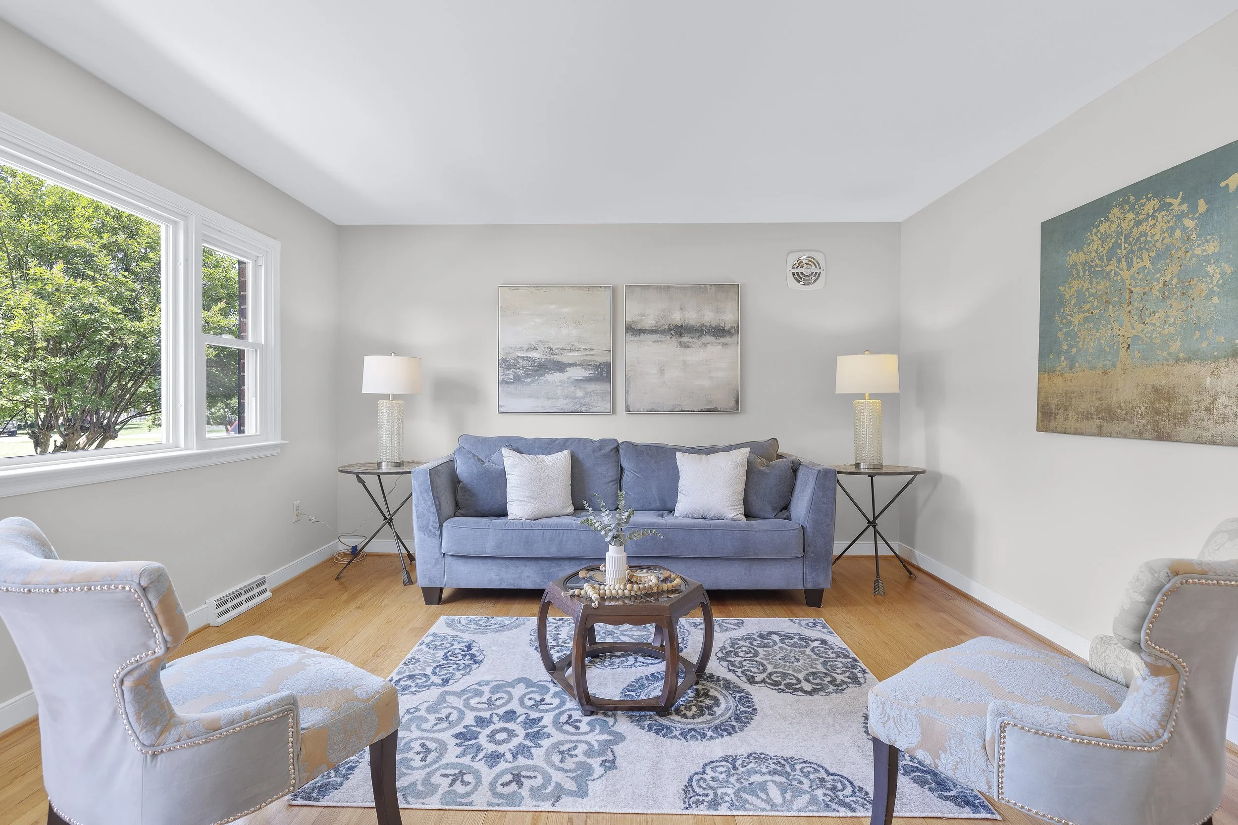A living room with a gray couch and armchairs, a round wooden coffee table with decorative beads, two side tables with lamps, two abstract art paintings on the wall, large window with greenery outside, hardwood floor, and blue patterned area rug.