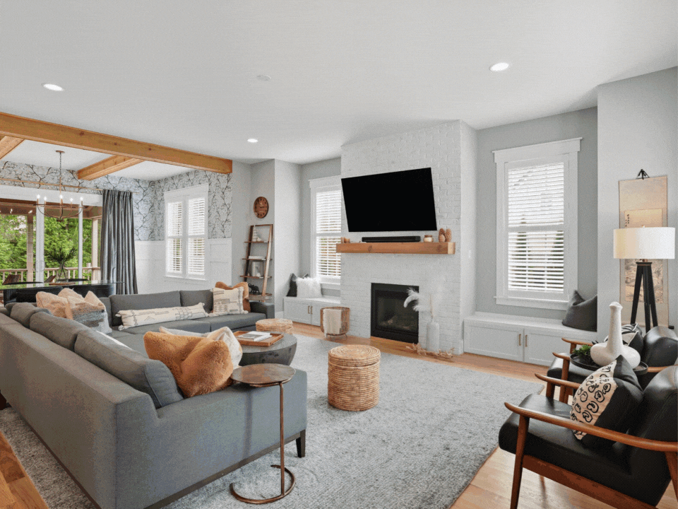 A cozy living room with gray couches, decorative pillows, a white brick fireplace, a mounted flat-screen TV, natural light from multiple windows, and a mix of modern and rustic decor.