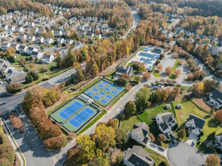 An aerial view of a suburban neighborhood with houses, roads, and a community center featuring multiple outdoor tennis courts with blue surfaces.