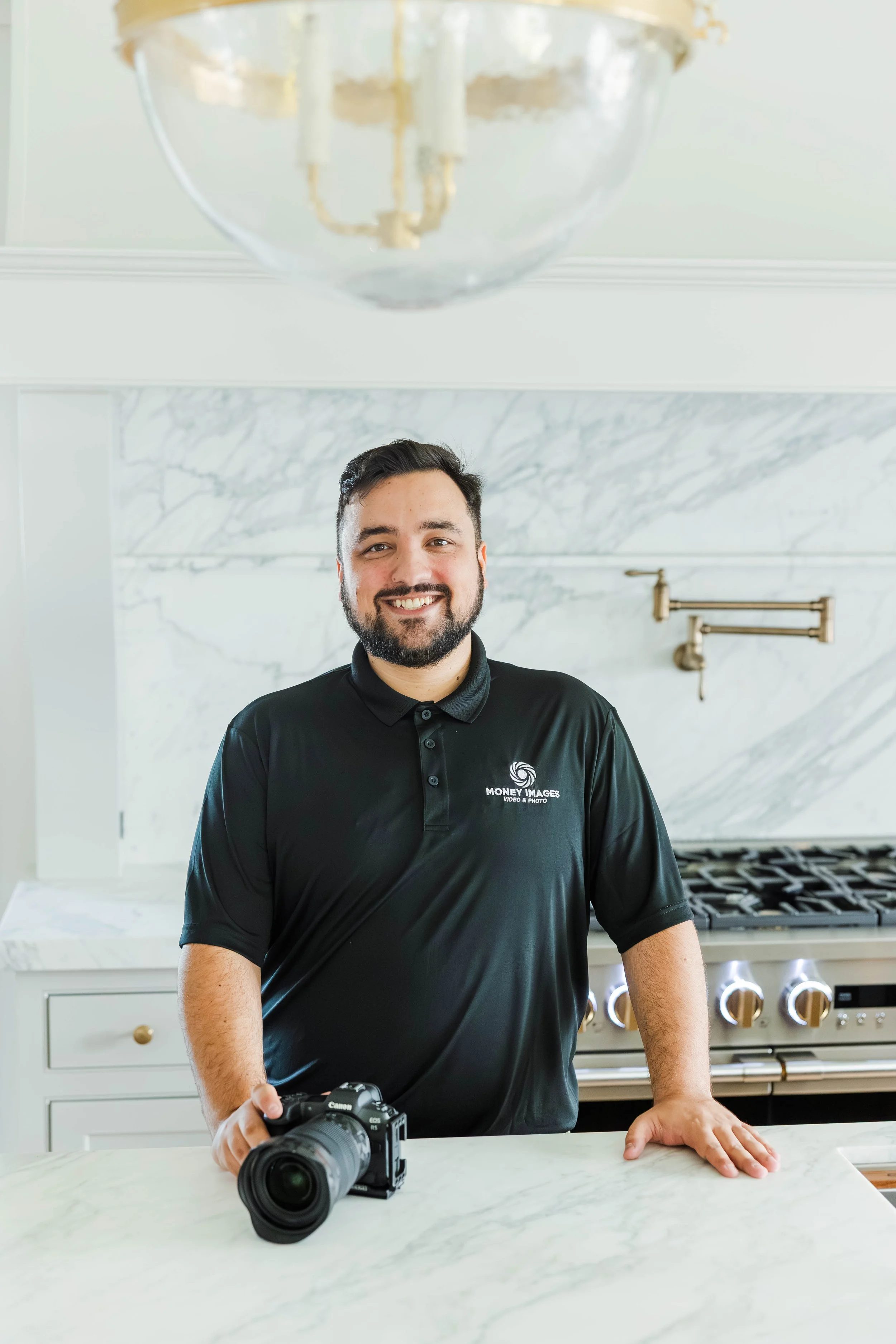 A man with a beard and dark hair smiling in a modern kitchen, wearing a black polo shirt with a logo, holding a camera in one hand and resting his other hand on a marble countertop, with a professional stove and marble backsplash in the background.