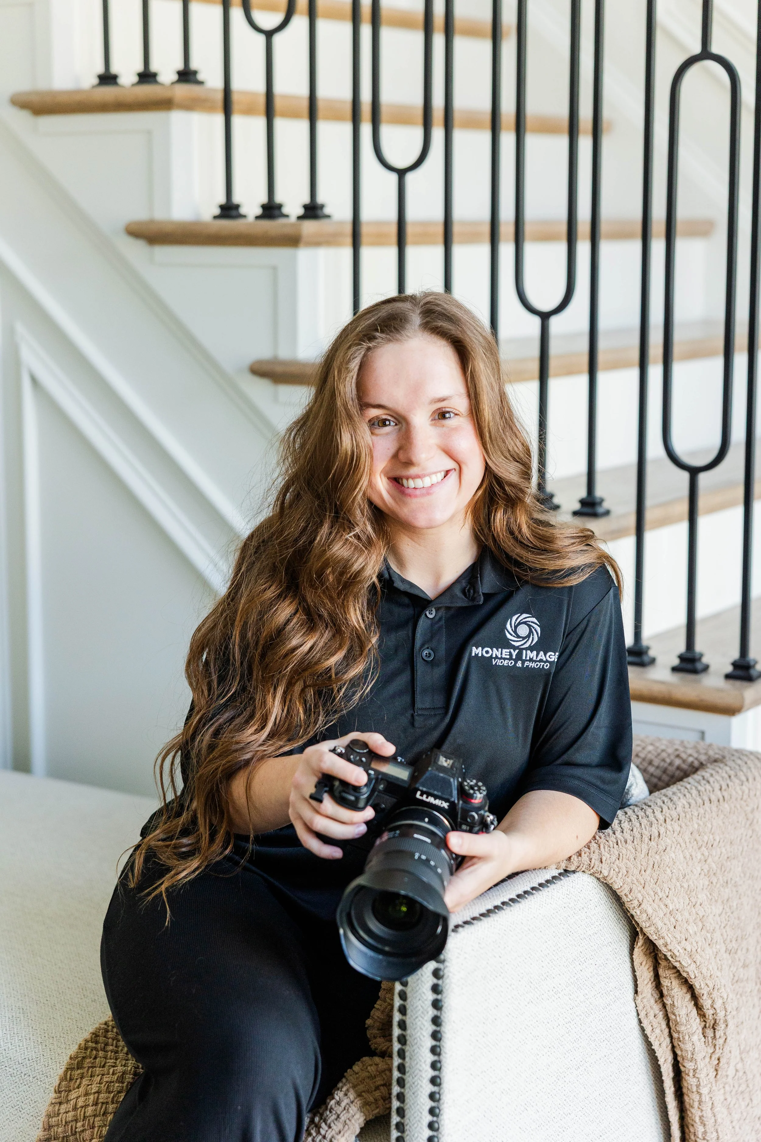 A woman with long wavy hair smiling while holding a professional camera, sitting on a beige couch with a textured brown blanket draped over the armrest, in a home interior with a staircase and black metal railings in the background.