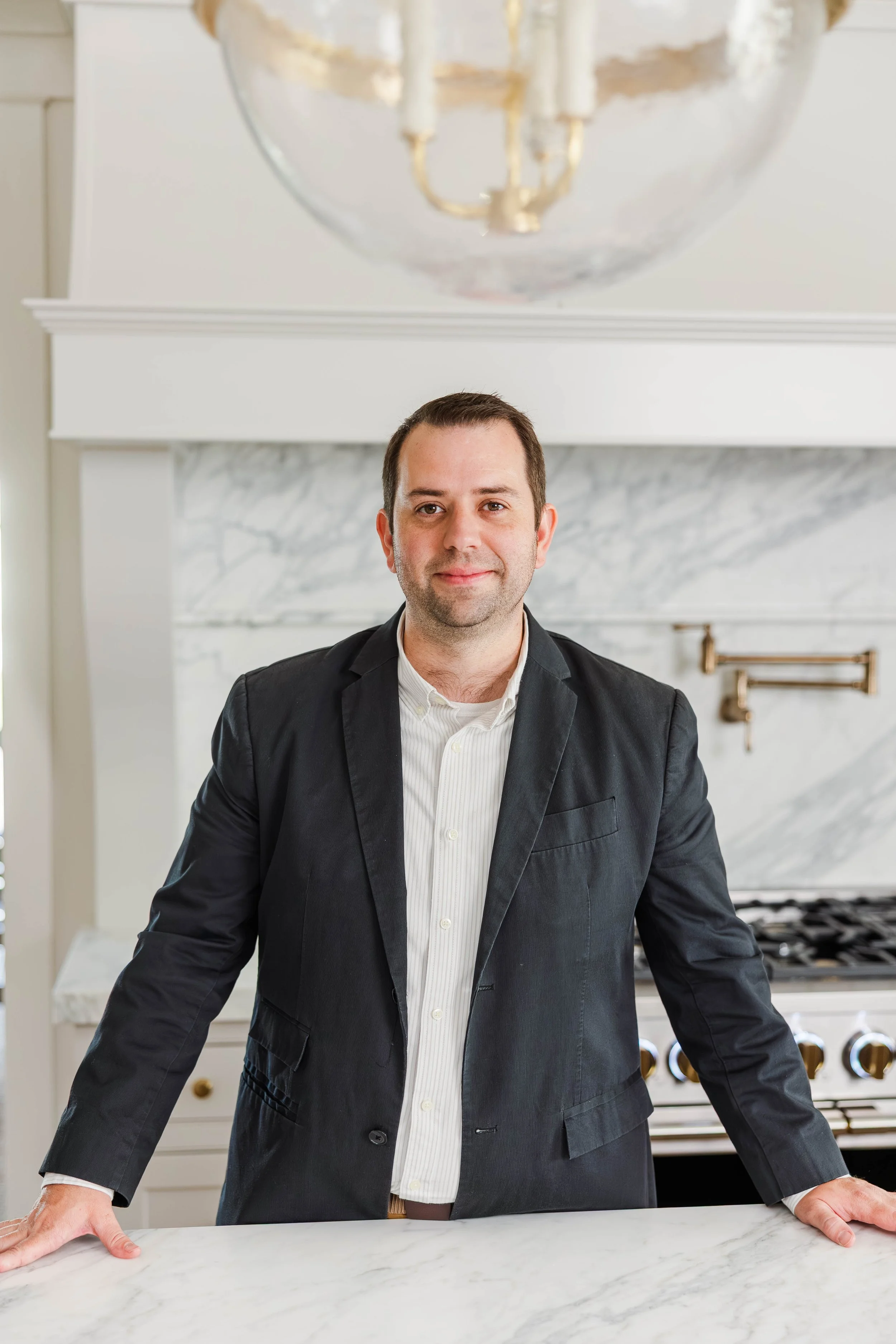A man in a black suit and white shirt leaning on a white marble kitchen island in a modern kitchen with a white marble backsplash, gas stove, and copper utensils hanging on the wall.