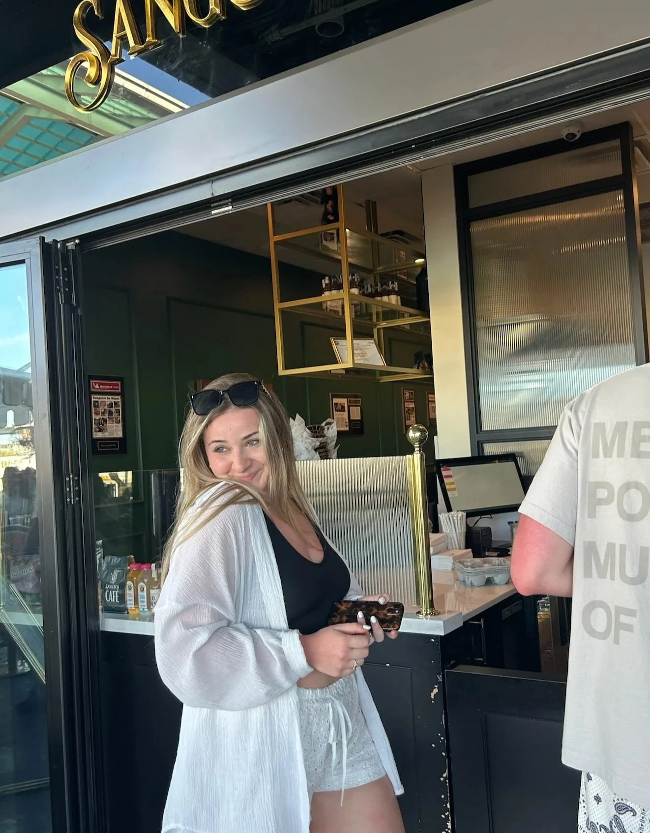 A young woman with long blonde hair, wearing sunglasses on her head, a black top, white shorts, and a white cardigan, smiling and holding a smartphone inside a coffee shop or cafe.