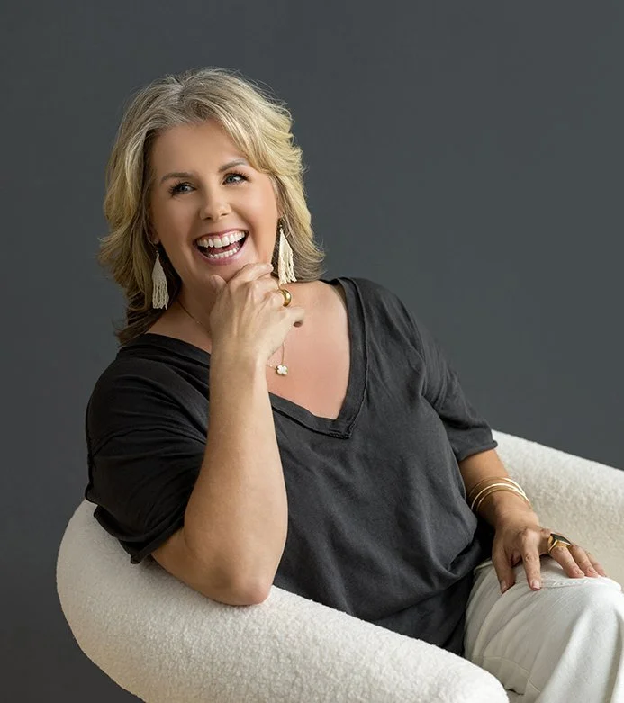 A woman with blond hair smiling and sitting in a white chair against a dark grey background, wearing a black top and jewelry.