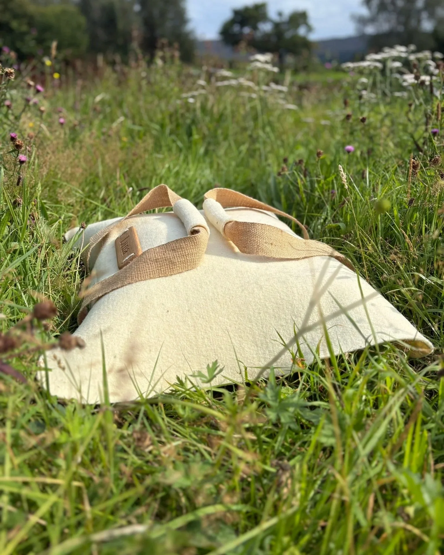 A creamy wool felt pet shroud with beige webbing handles on the grass in a field with wildflowers, trees and a blue sky in the background.