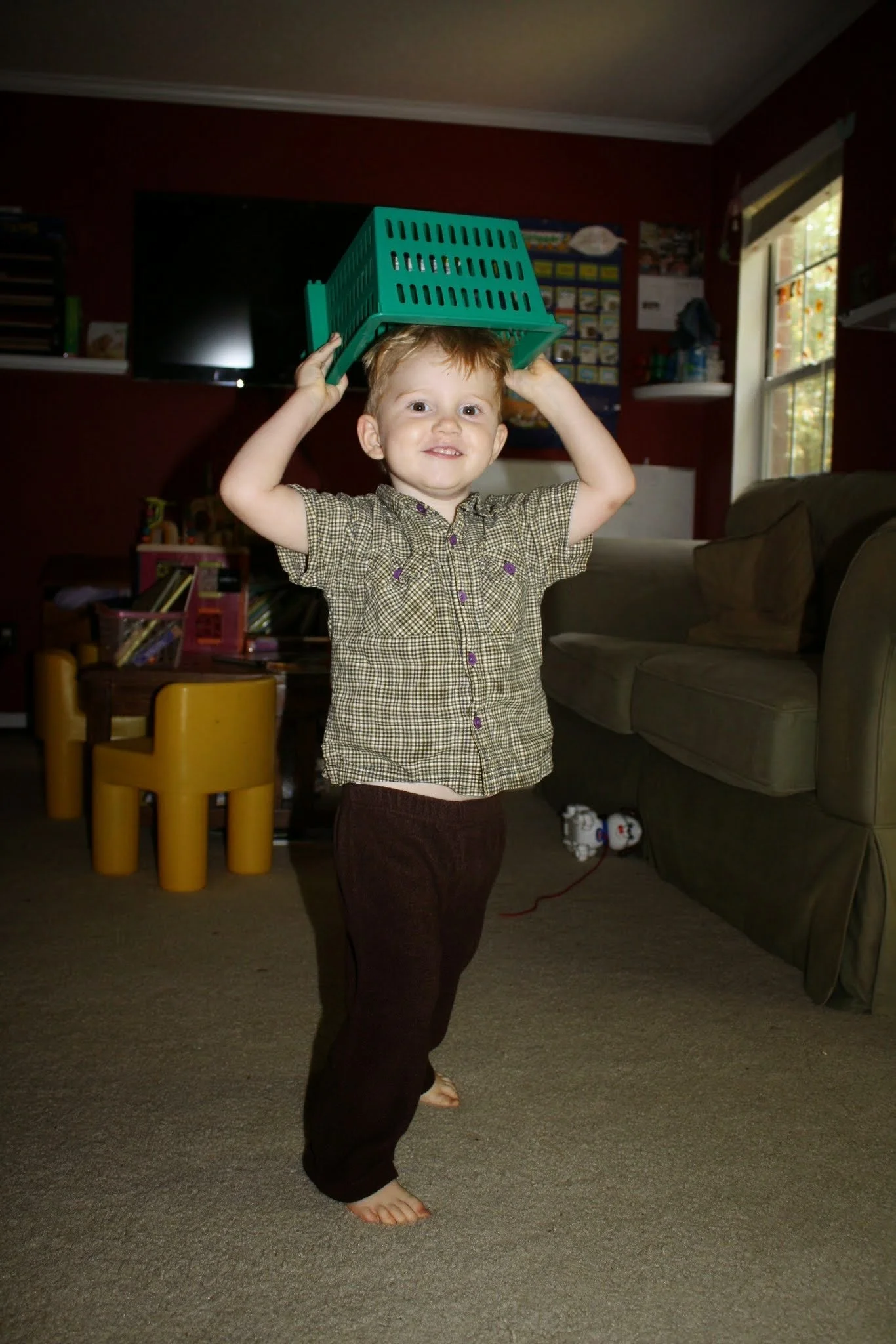 Smiling toddler balancing a green basket on his head during playful indoor activity, representing play-based pediatric speech therapy services at Play to Say Therapy