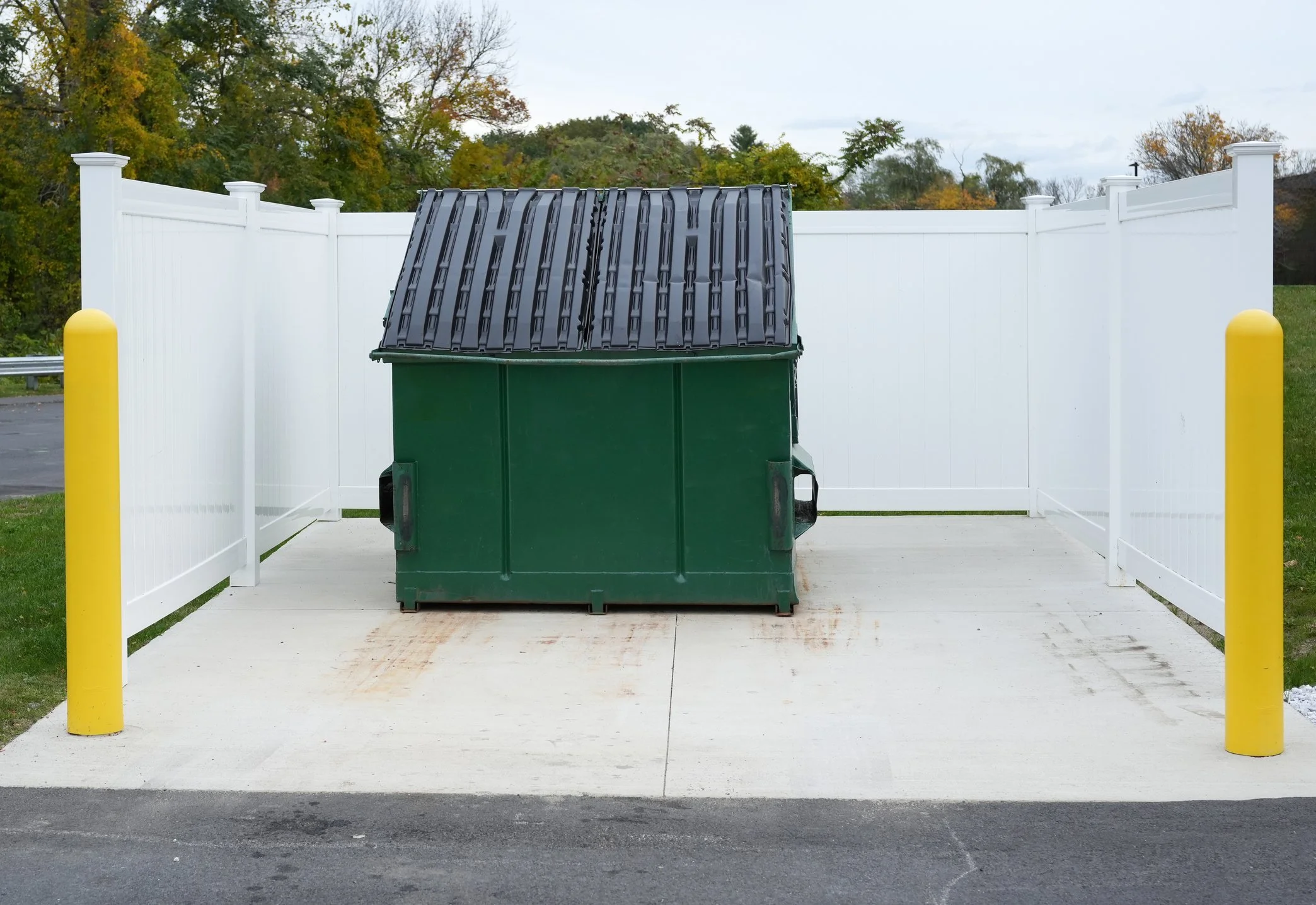 A green trash bin with a black lid is situated on a white platform in front of a white fence, with yellow posts on either side. The background features trees with fall foliage and a cloudy sky.