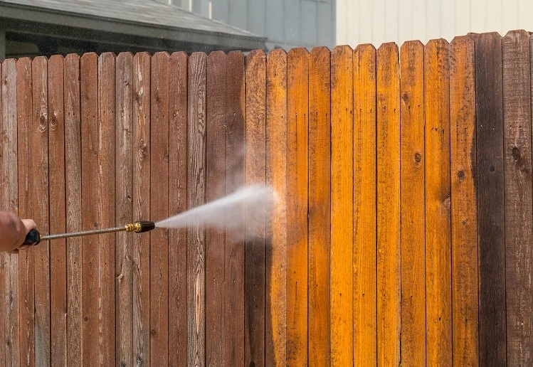 Person using a power washer to remove dirt or grime from a wooden fence, creating a spray of water.
