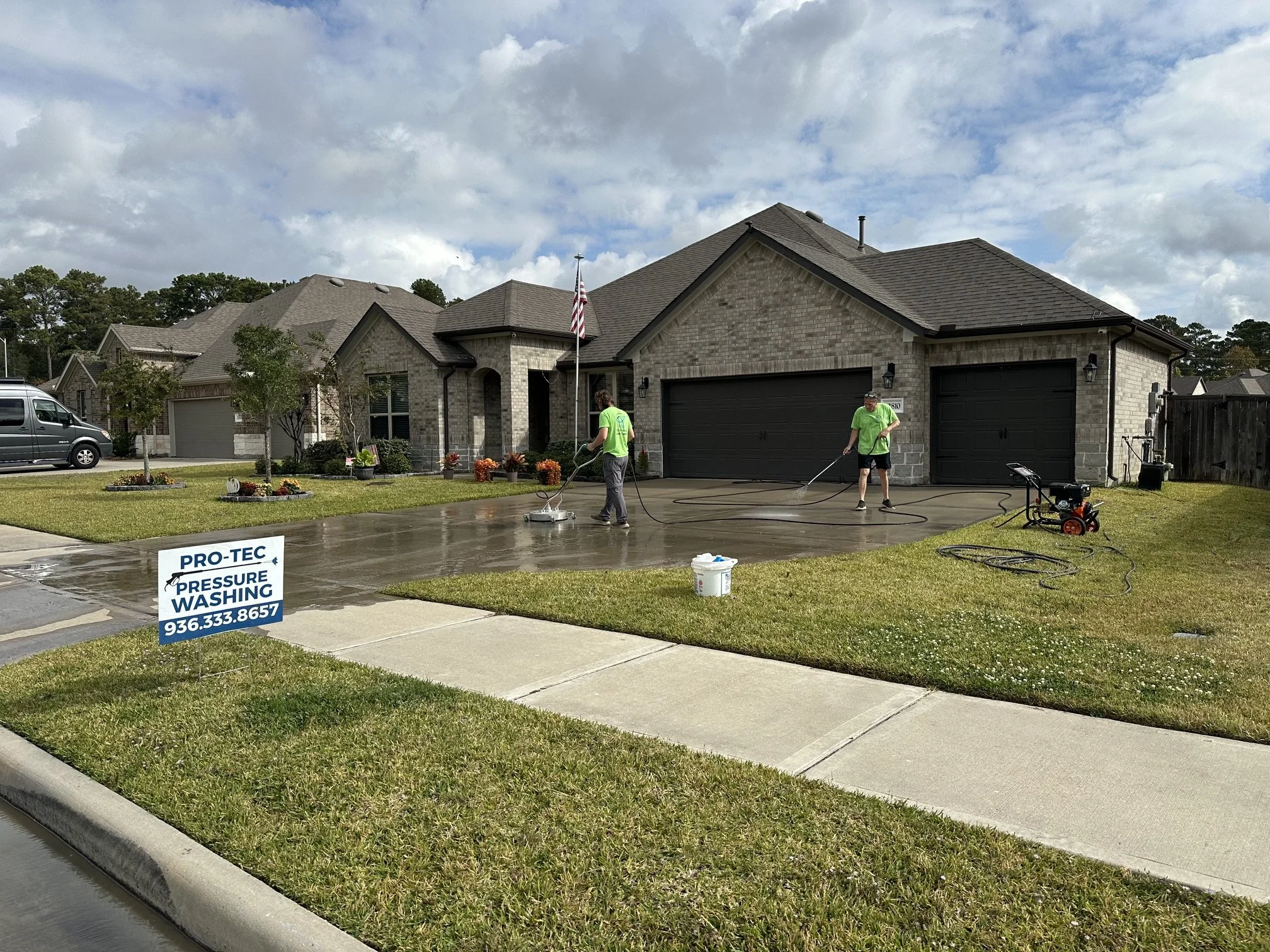 Two workers pressure washing a concrete driveway in front of a suburban house with a sign reading 'PRO-TEC Pressure Washing' and a phone number, on a cloudy day.