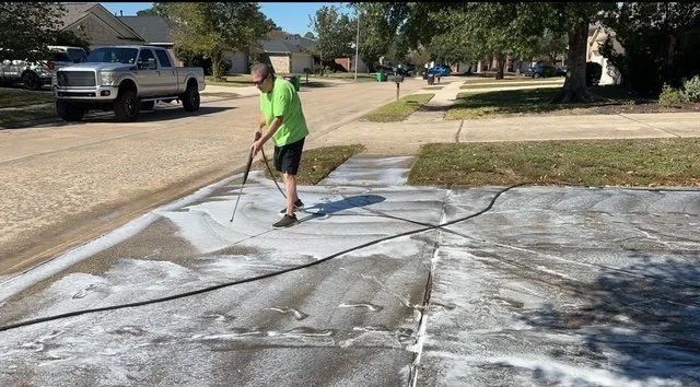 A person wearing a bright green shirt and black shorts power washing a concrete driveway on a residential street.