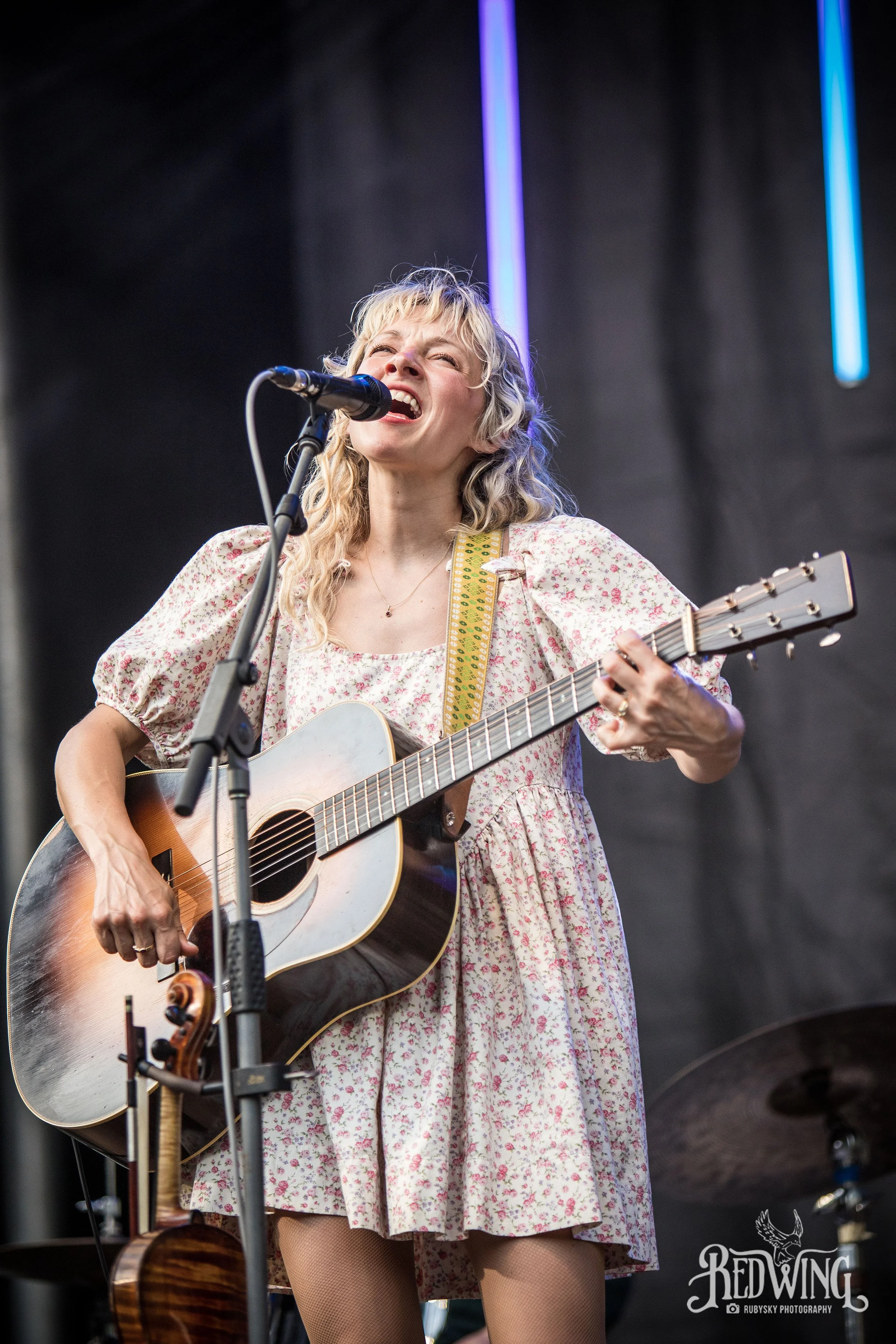 A woman with curly blonde hair singing passionately into a microphone and playing an acoustic guitar during a live outdoor performance.