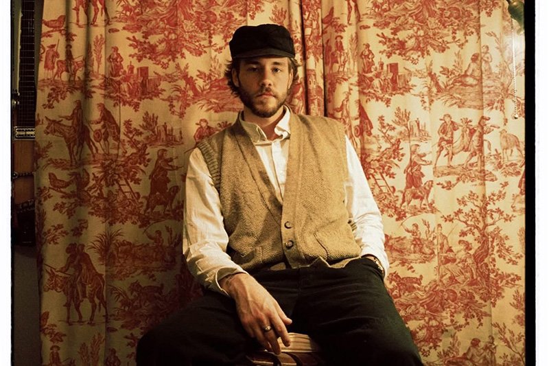Young man with a beard wearing a black cap, white shirt, and beige vest, sitting in front of a vintage red and beige patterned curtain.