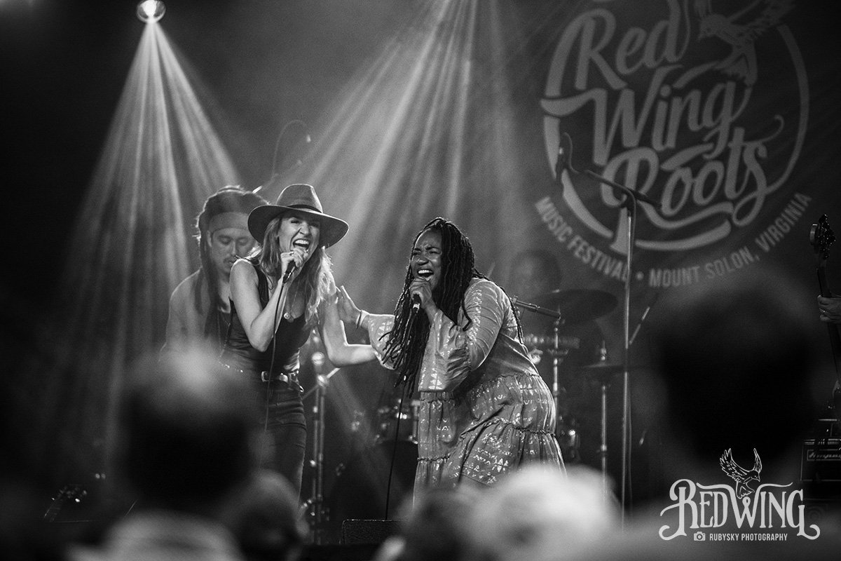 Two women singing passionately on stage at the Red Wing Roots music festival, with bright stage lights shining down on them. One woman is wearing a wide-brimmed hat, and both appear to be enjoying the performance. There are musical instruments and a microphone stand visible, with a crowd in the foreground and a festival sign in the background.