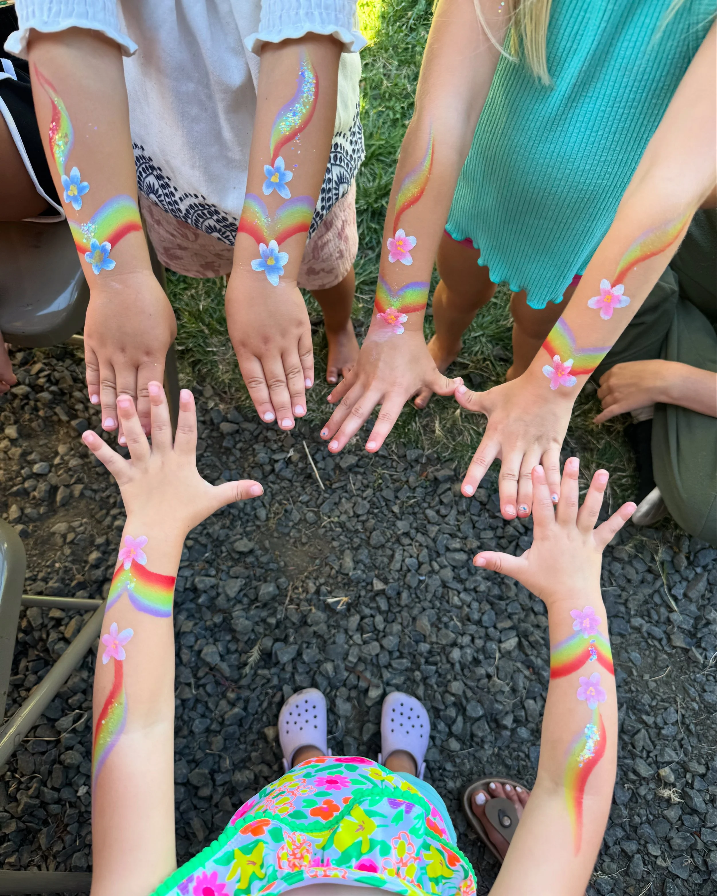 Children with rainbow and flower face painting and arm painting, standing outdoors at a little kids party in Eugene, Oregon, showing their painted arms and legs from above.