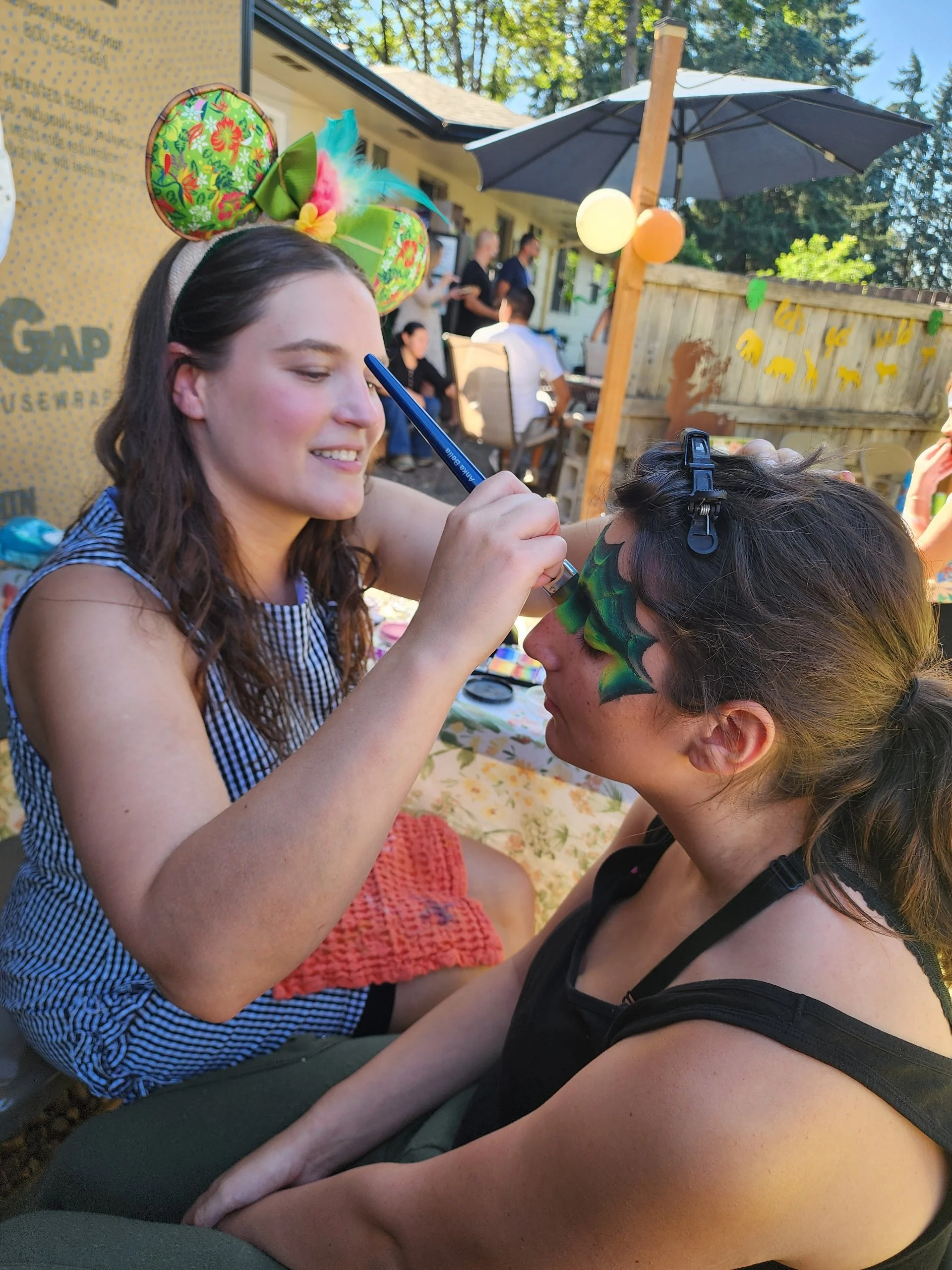 A young woman wearing a colorful headband with a bow and feathers paints a mermaid face painting design on a young girl's face, seated outdoors at a backyard  party with balloons and people in the background.