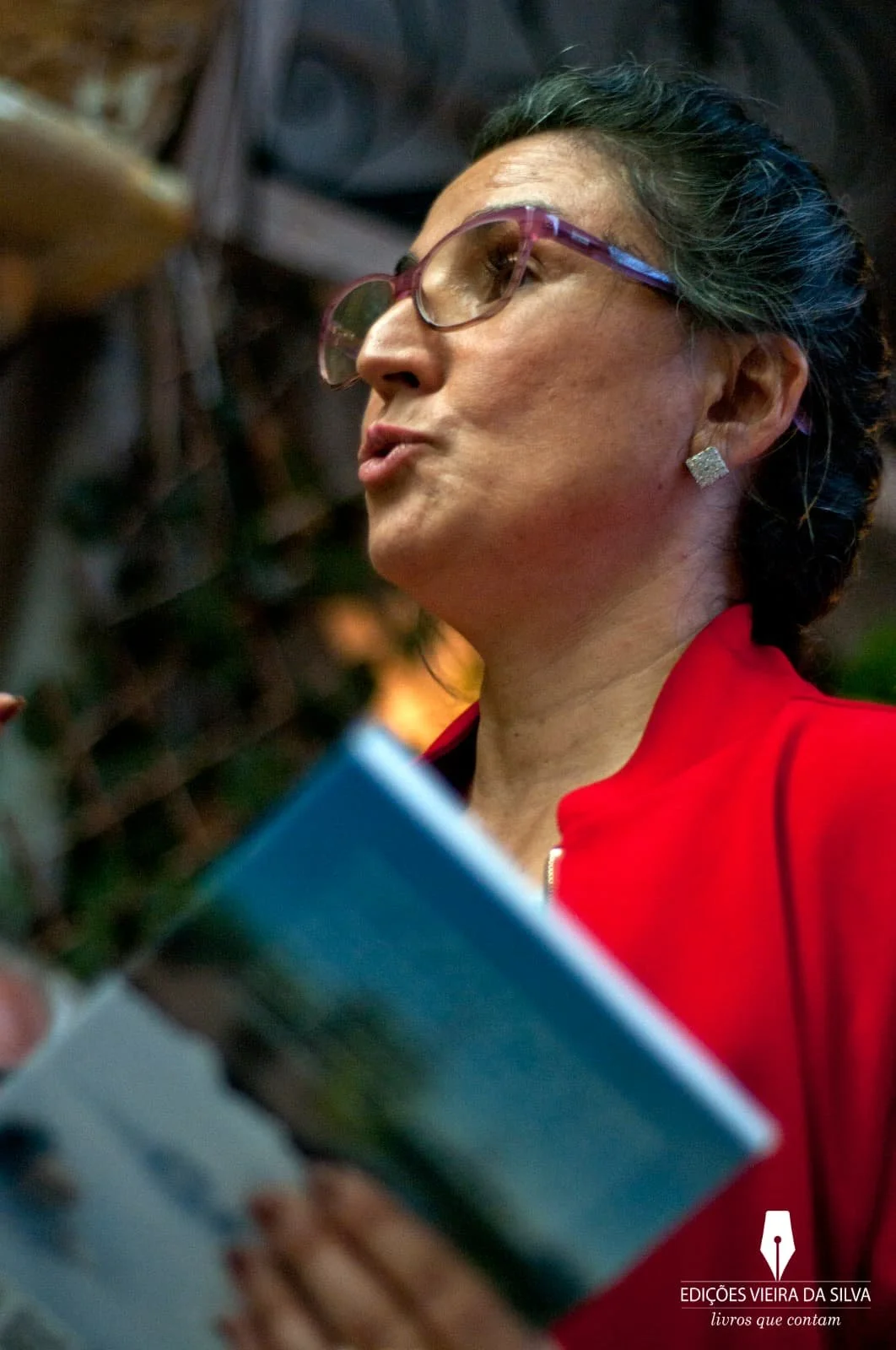 A woman with gray and black hair styled in a bun, wearing glasses and diamond earrings, is holding a book or magazine while speaking or reading aloud. She is dressed in a red jacket.