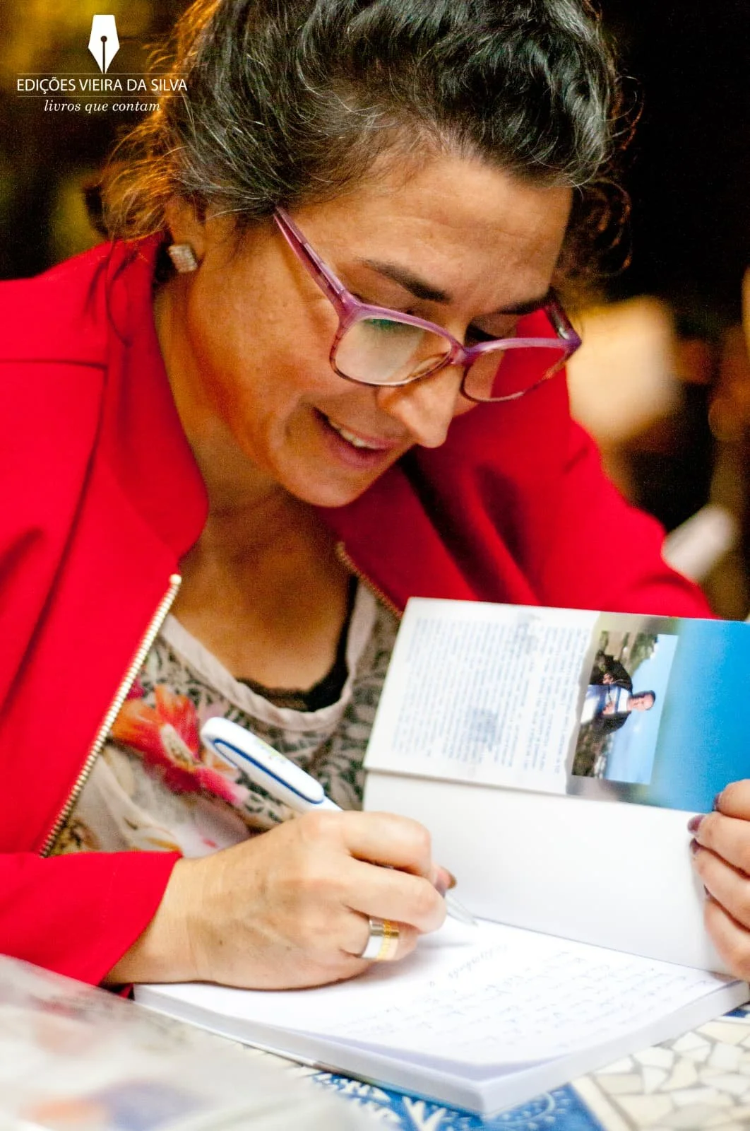 A woman wearing glasses and a red jacket is smiling while writing in a notebook. She is looking at a magazine or brochure that features a photograph of a person sitting by the seaside.