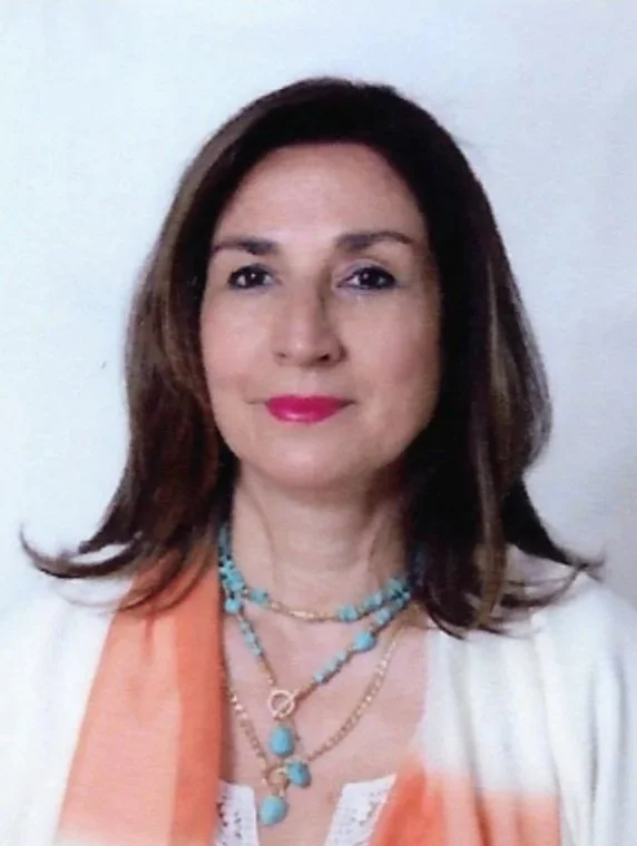 A woman with shoulder-length dark brown hair, wearing a white top with an orange scarf, and multiple turquoise and gold necklaces, standing against a plain white wall.