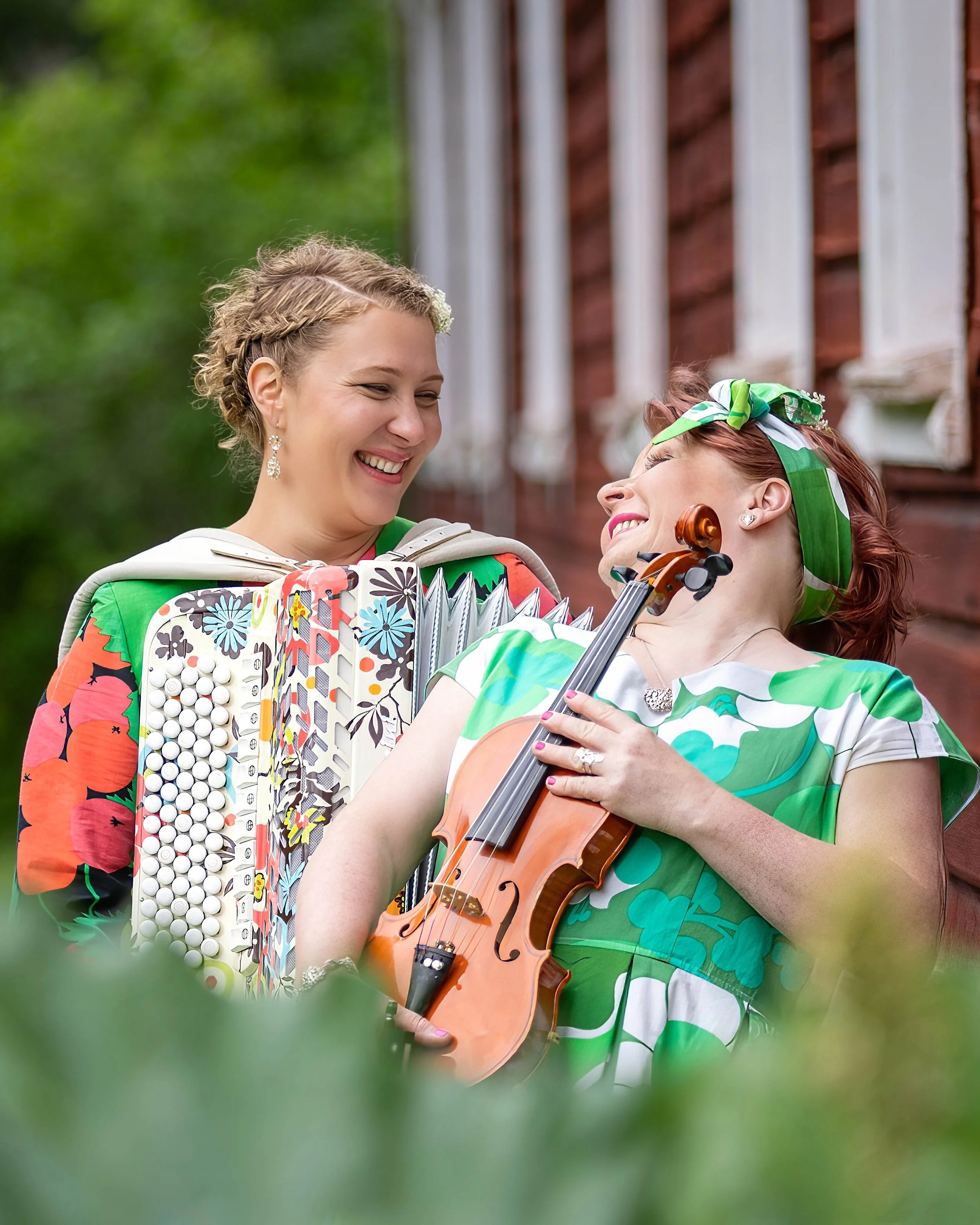 Kaksi muusikkoa esiintyy ulkona iloisesti viulun ja harmonikan säestyksellä. Punainen hirsitalo ja vehreä kasvillisuus luovat luonnonläheisen ja kutsuvan tunnelman.