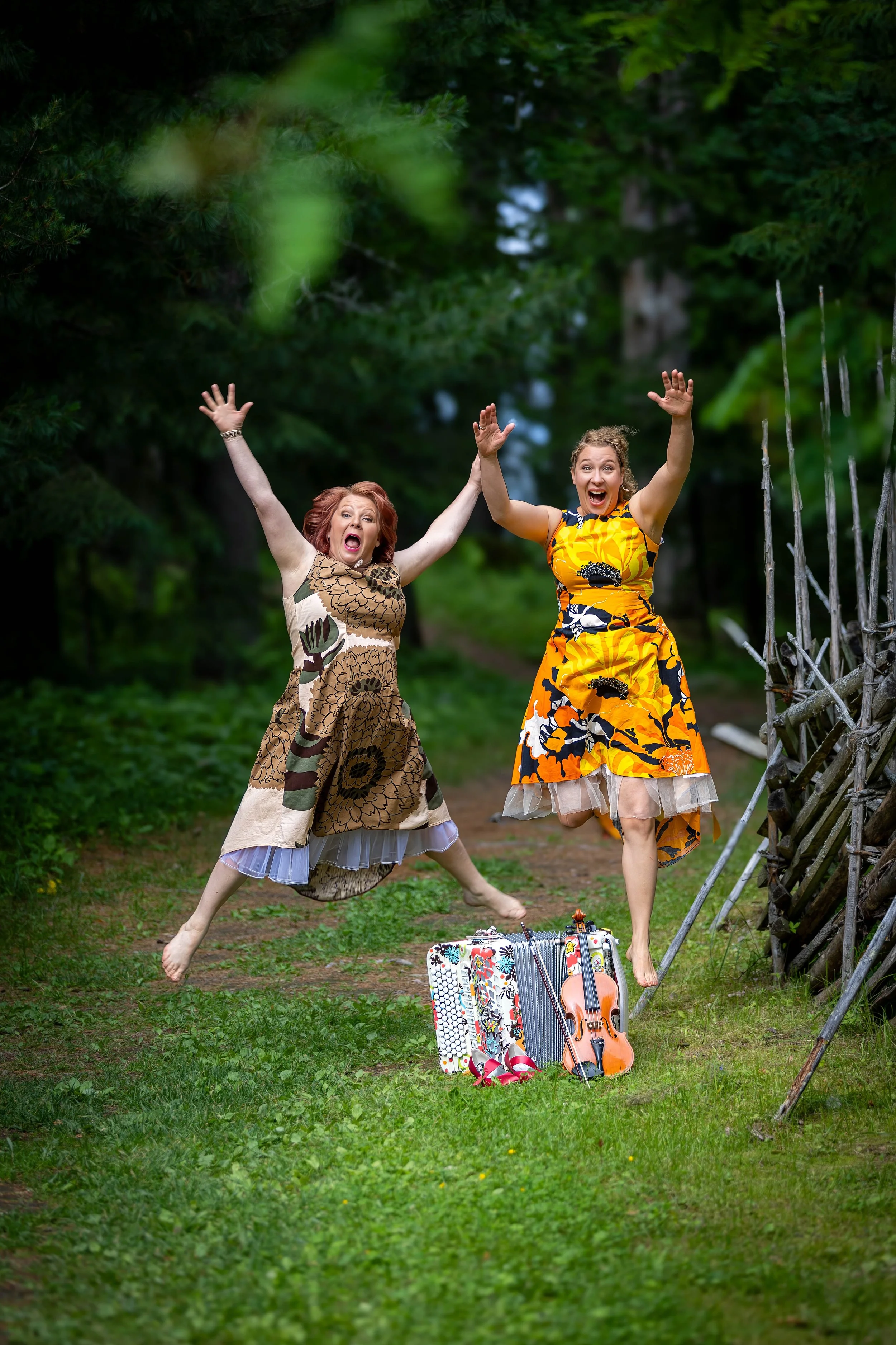 Riitta ja Petra hyppäävät iloisesti metsäpolulla viulun ja harmonikan kanssa. Taustalla kohoavat puut luovat kuvalle luonnonläheisen ja energisen tunnelman.