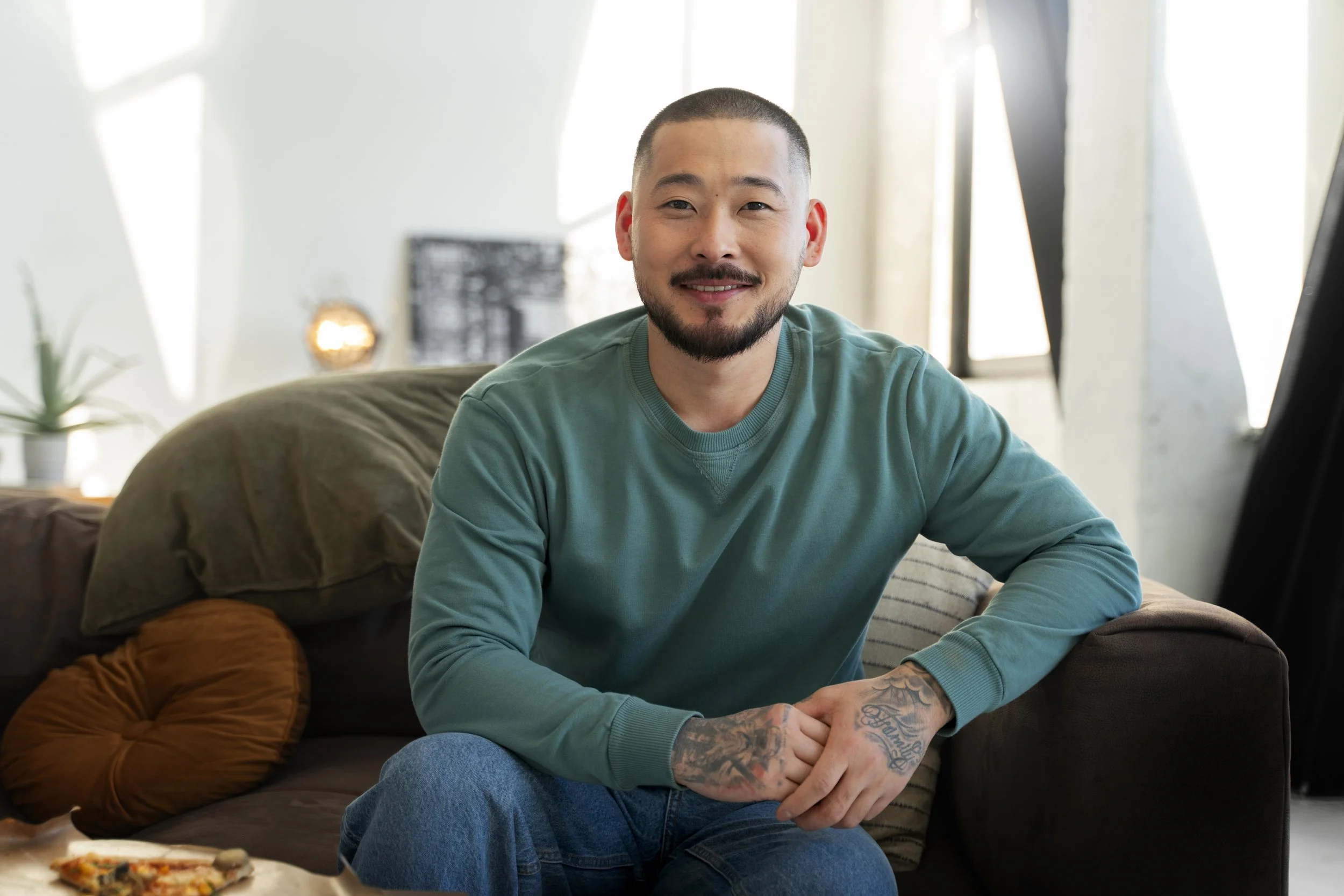 A young man with a beard and tattoos on his hands, sitting on a sofa in a bright, modern living room, smiling at the camera.