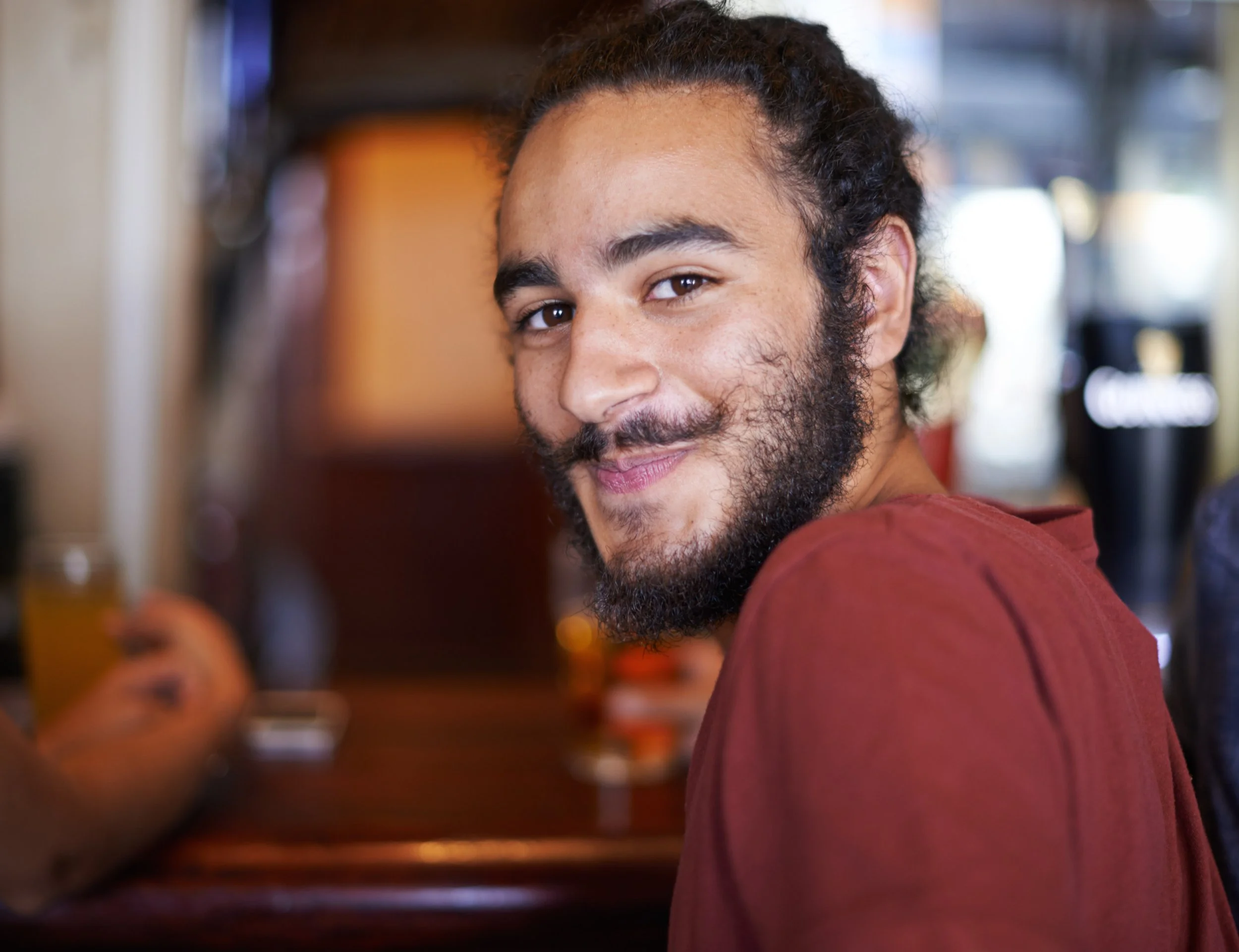 A smiling man with curly hair and a beard, wearing a red shirt, sitting at a table in a restaurant or cafe.