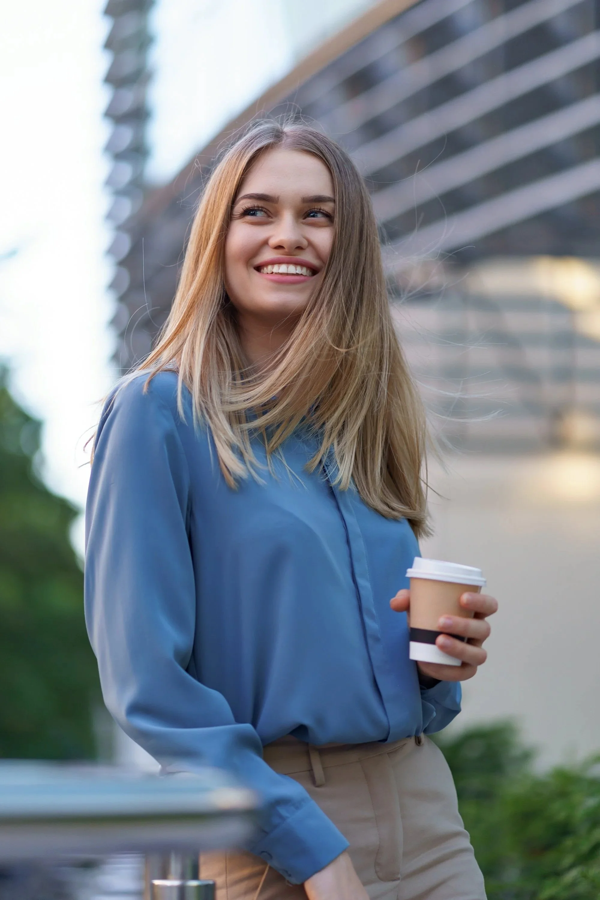 Young woman with long blond hair smiling outdoors, holding a coffee cup, wearing a blue blouse and beige pants.