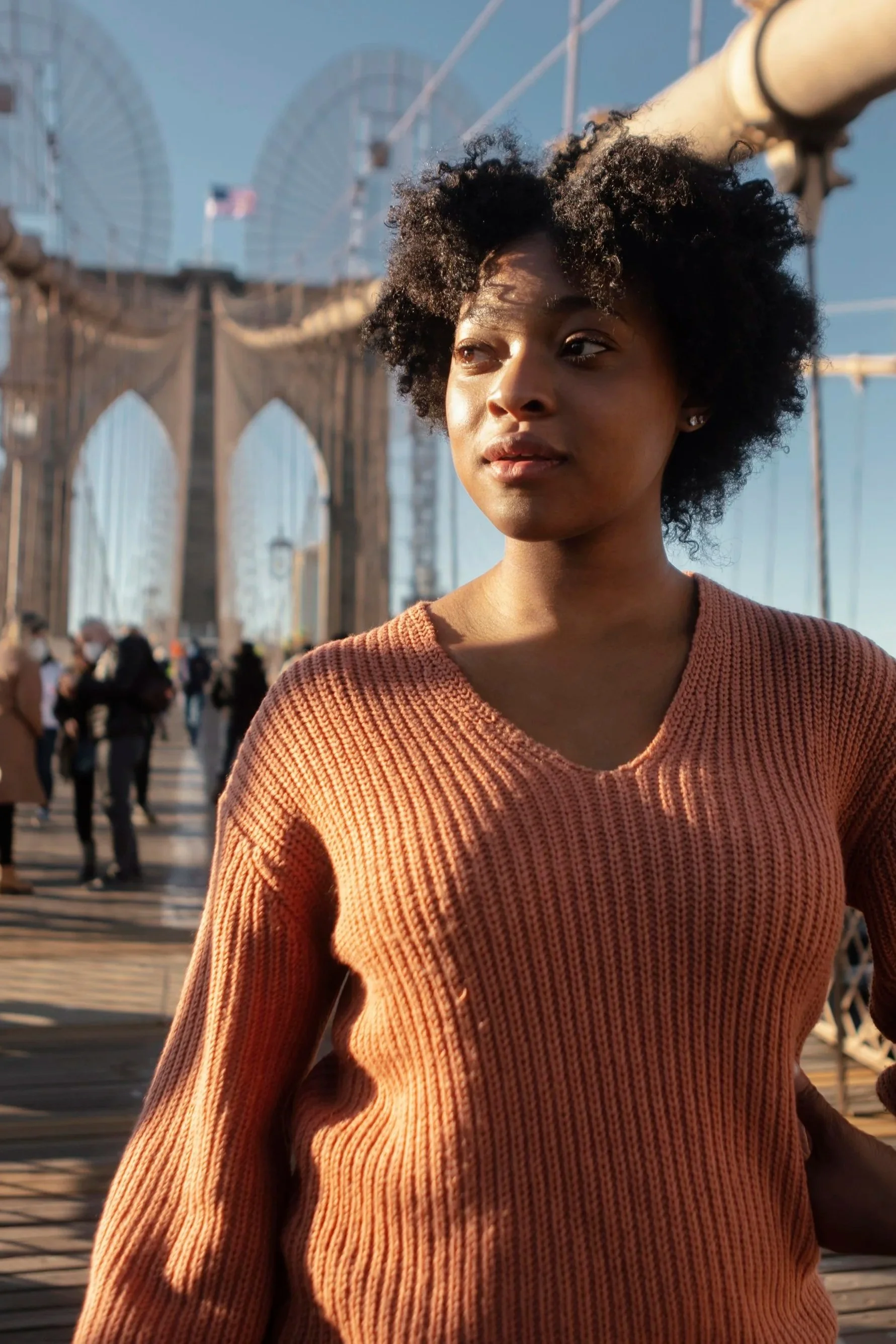 A woman standing on the Brooklyn Bridge with the New York City skyline in the background, wearing a rust-colored sweater and looking into the distance.